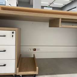 Electro4you - Biurko z szufladami i gniazdkami elektrycznymi w listwie przypodłogowej. Widoczne gniazdka z bolcem i bez, oraz włączniki. Blat biurka z jasnego drewna.