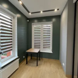 Nowoczesne wnętrze z oświetleniem punktowym w suficie, zieloną ścianą z wzorem, oknami z żaluzjami i małym stolikiem. Widoczny grzejnik i fragment instalacji elektrycznej.
