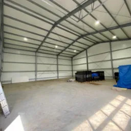 Wnętrze hali z metalową konstrukcją i oświetleniem LED. Widoczne materiały budowlane i drabina oparta o ścianę. Jasne, dzienne światło.