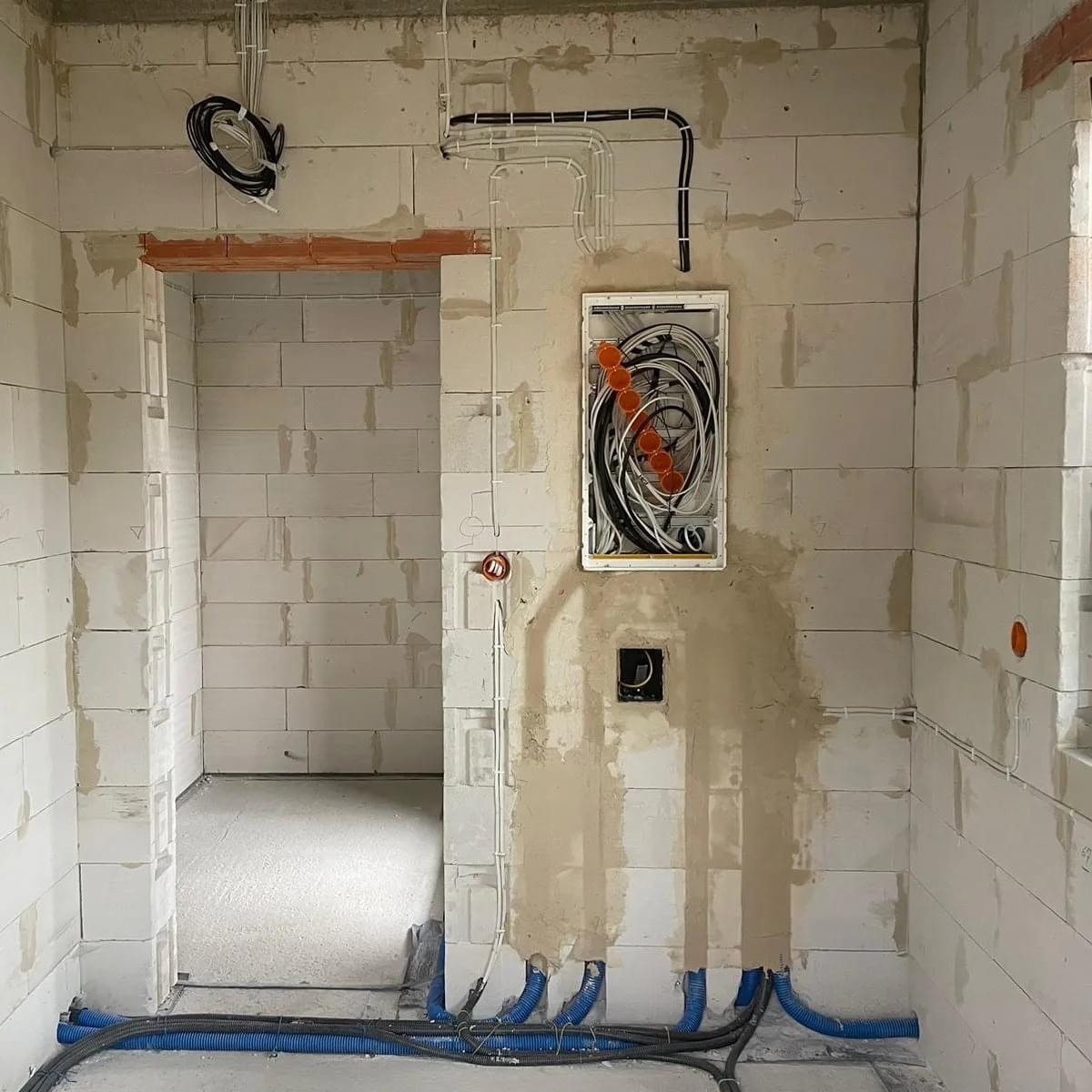 Wnętrze w trakcie prac elektrycznych: widoczne kable prowadzone po ścianach z białych bloczków, puszka elektryczna z pomarańczowymi złączkami, oraz niebieskie rury osłonowe przy podłodze. Ściany...