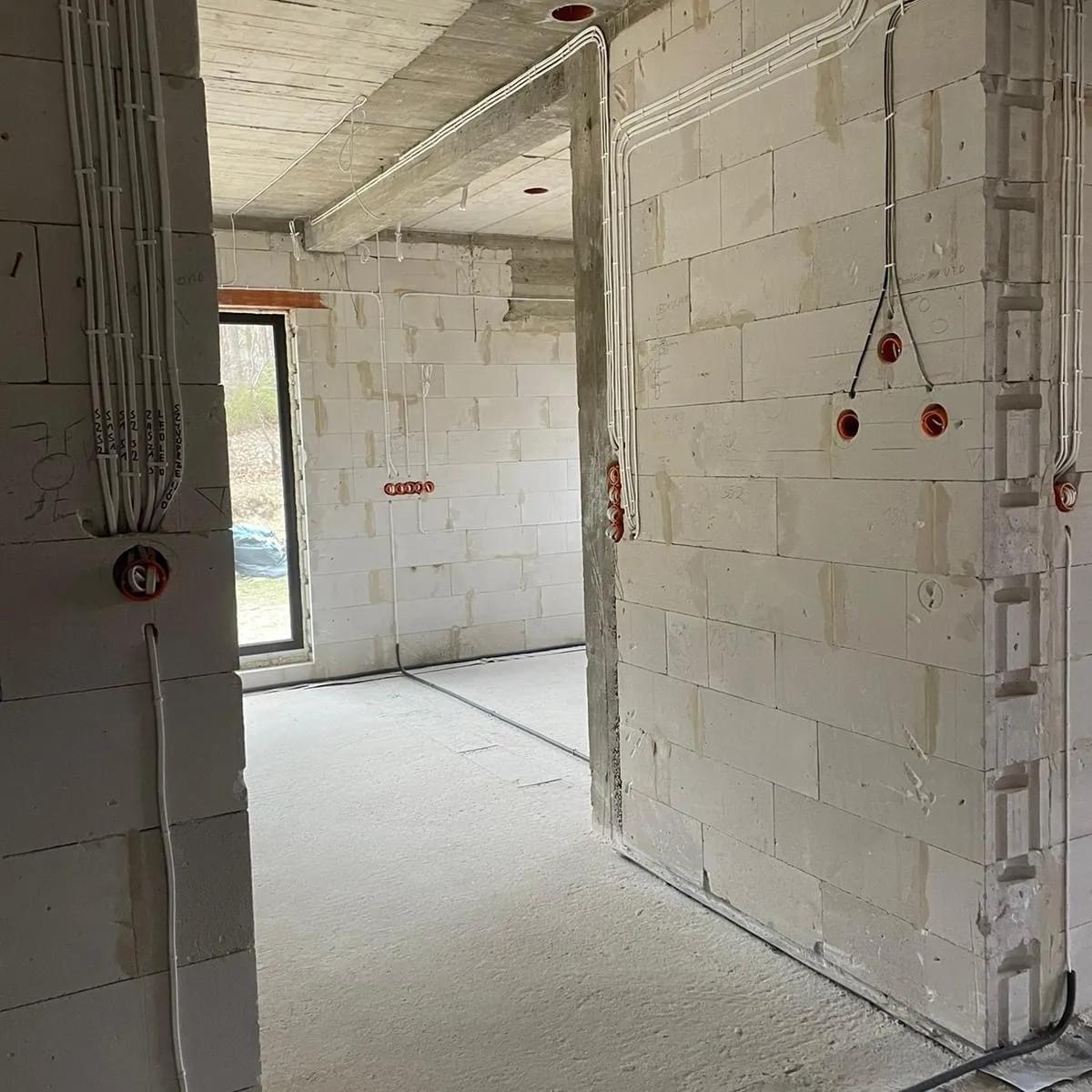 Surowy stan wnętrza budynku w trakcie prac elektrycznych: białe ściany z betonu komórkowego, widoczne peszle z przewodami elektrycznymi ułożone natynkowo, puszki instalacyjne w kolorze...