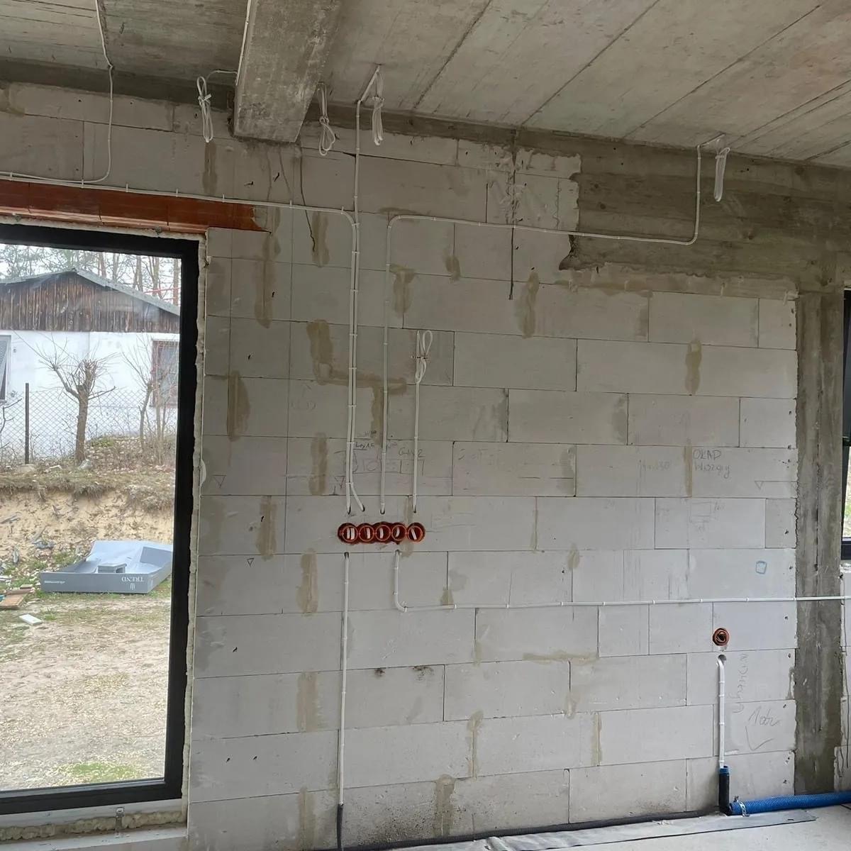 Wnętrze w stanie surowym z widoczną instalacją elektryczną: kable prowadzone po ścianie z betonu komórkowego, czerwone puszki pod gniazdka, kable zwisające z sufitu, widoczne zapiski na ścianie.
