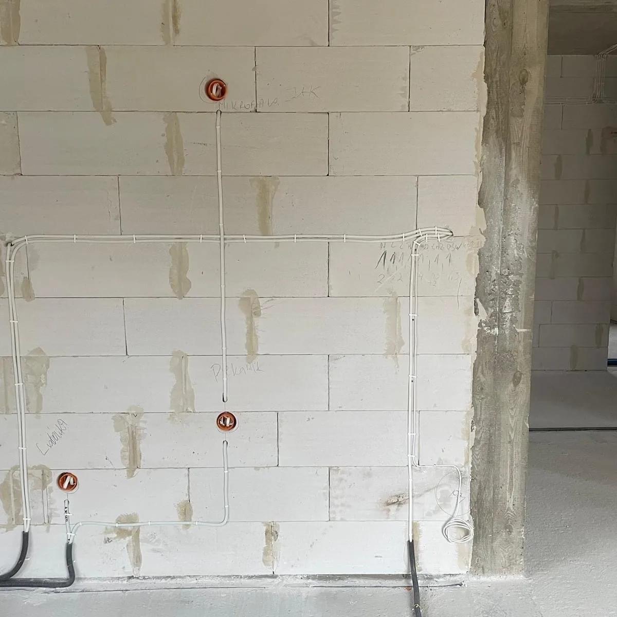 Ściana z betonu komórkowego w trakcie budowy z poprowadzonymi przewodami elektrycznymi w białych rurkach, widoczne puszki instalacyjne i odręczne notatki na ścianie.