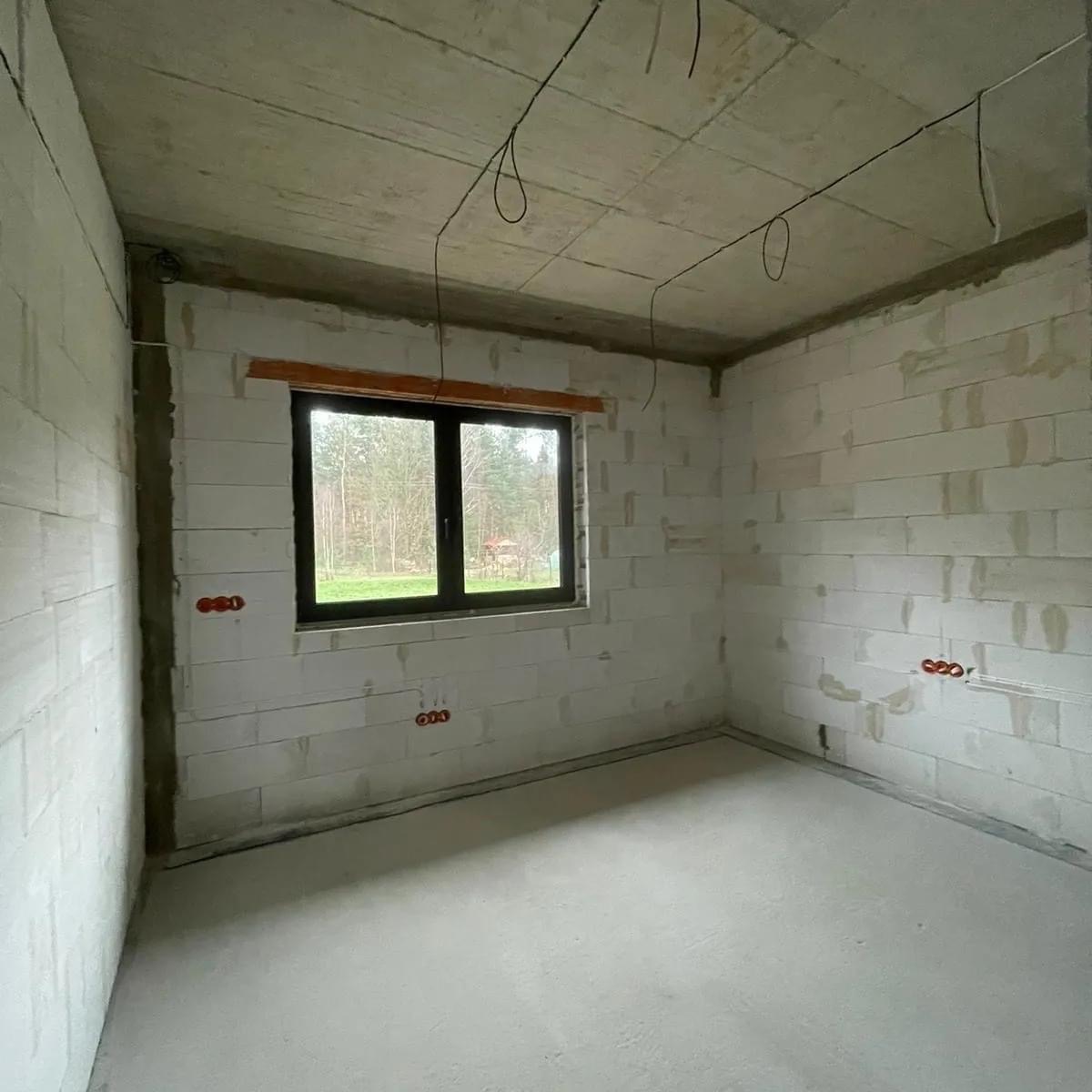 Surowy pokój w trakcie wykończenia: ściany z białego bloczka, wylewka betonowa, okno z widokiem na las i wystające kable elektryczne z sufitu oraz wyprowadzone gniazdka w ścianach.