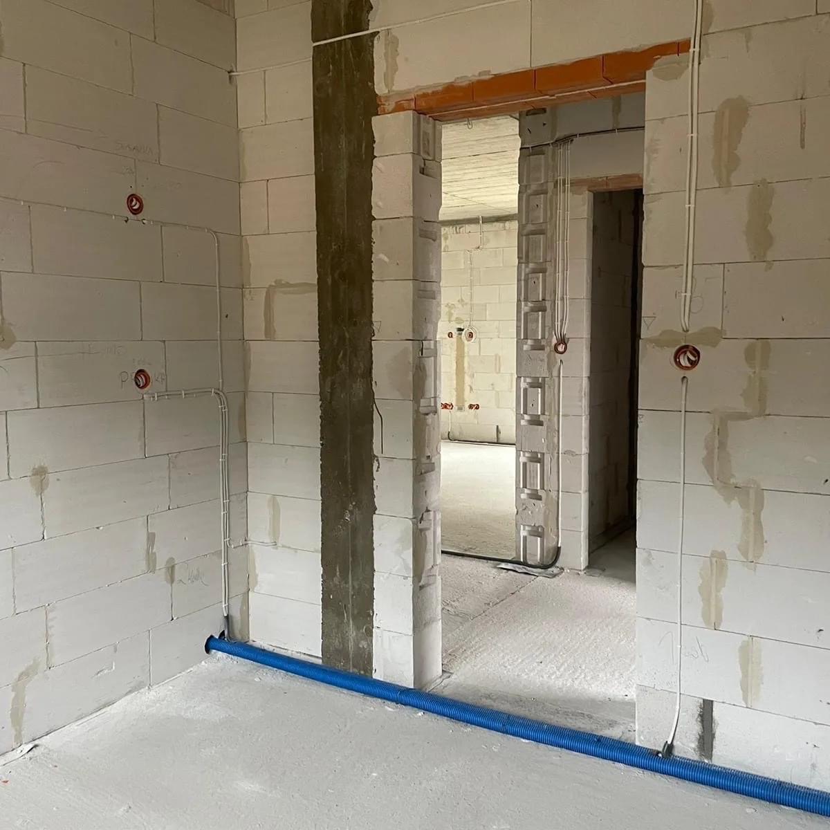 Wnętrze w stanie surowym z widoczną instalacją elektryczną: przewody w ścianach z betonu komórkowego oraz niebieska rura karbowana przy podłodze.