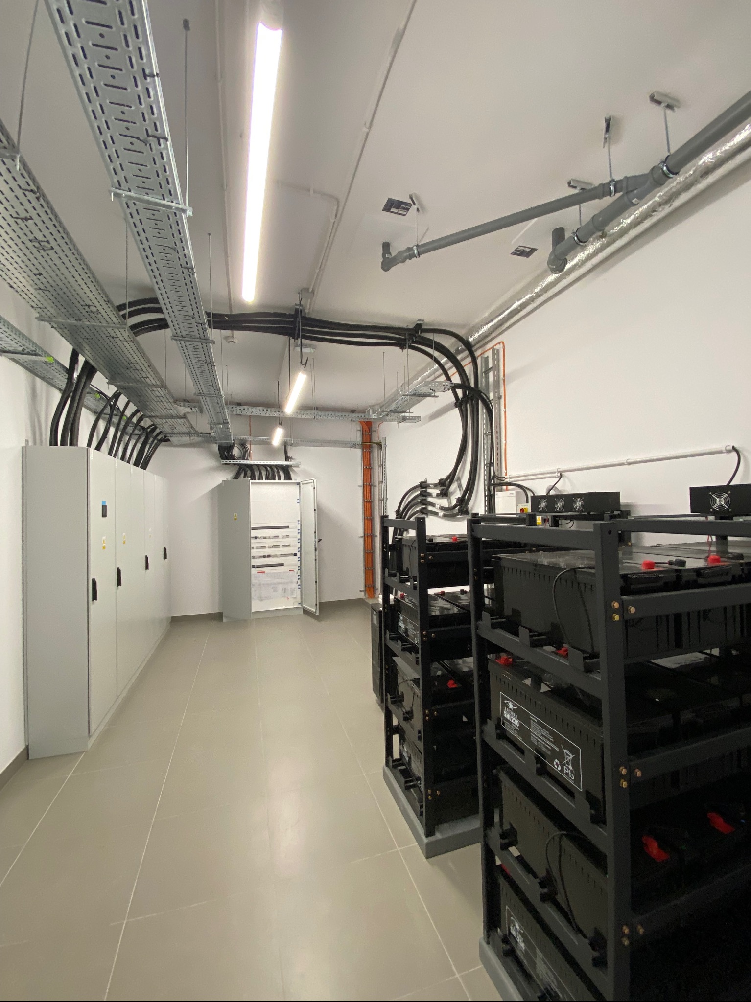 Wnętrze pomieszczenia technicznego w Klwowie z szafami elektrycznymi, rozdzielnicą z otwartymi drzwiami, korytami kablowymi pod sufitem oraz regałami z akumulatorami.