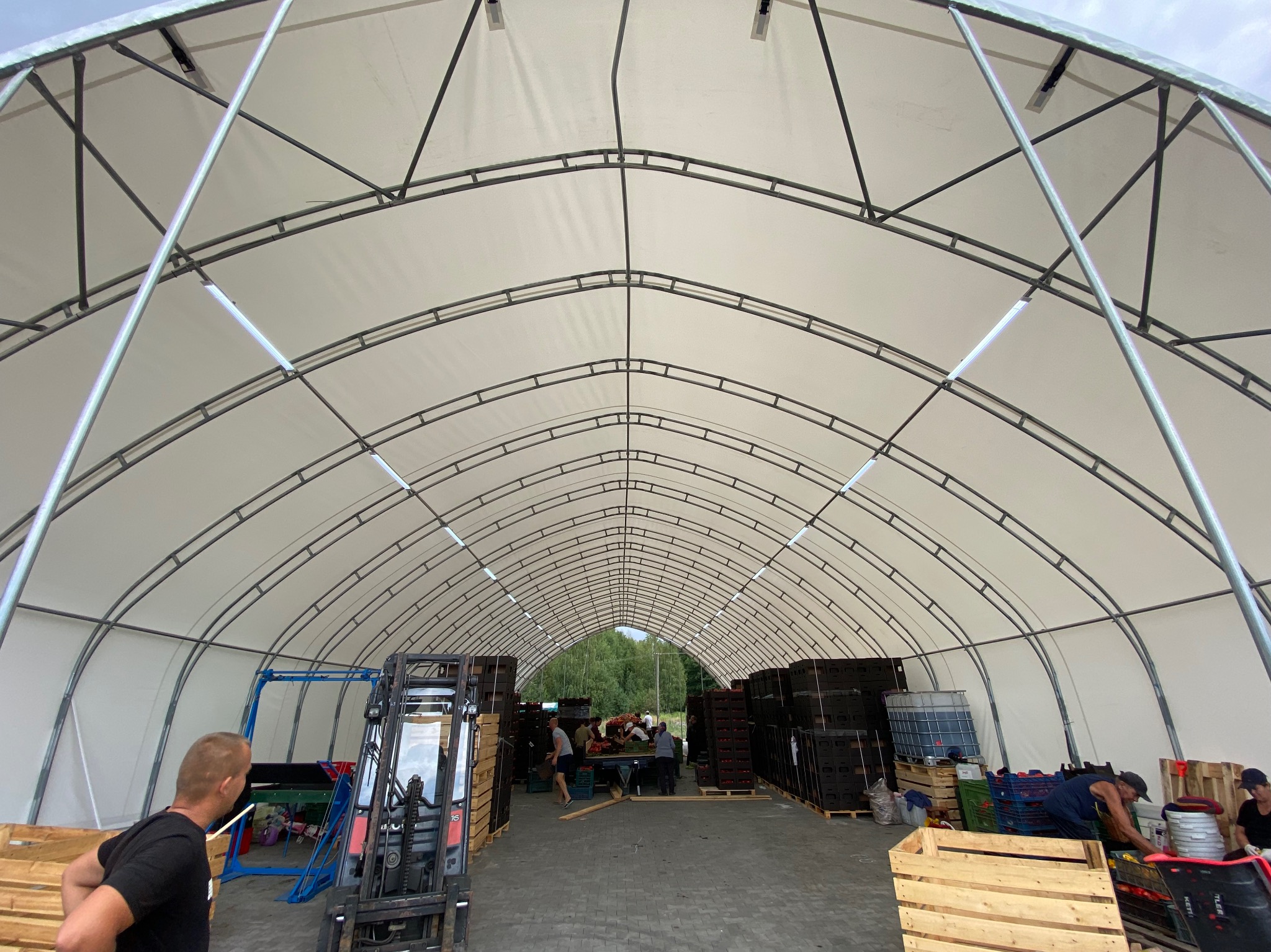 Wnętrze dużego, namiotowego magazynu z metalową konstrukcją i białym, materiałowym dachem, oświetlonego lampami LED, z widocznym wózkiem widłowym, paletami i osobami pracującymi w tle.