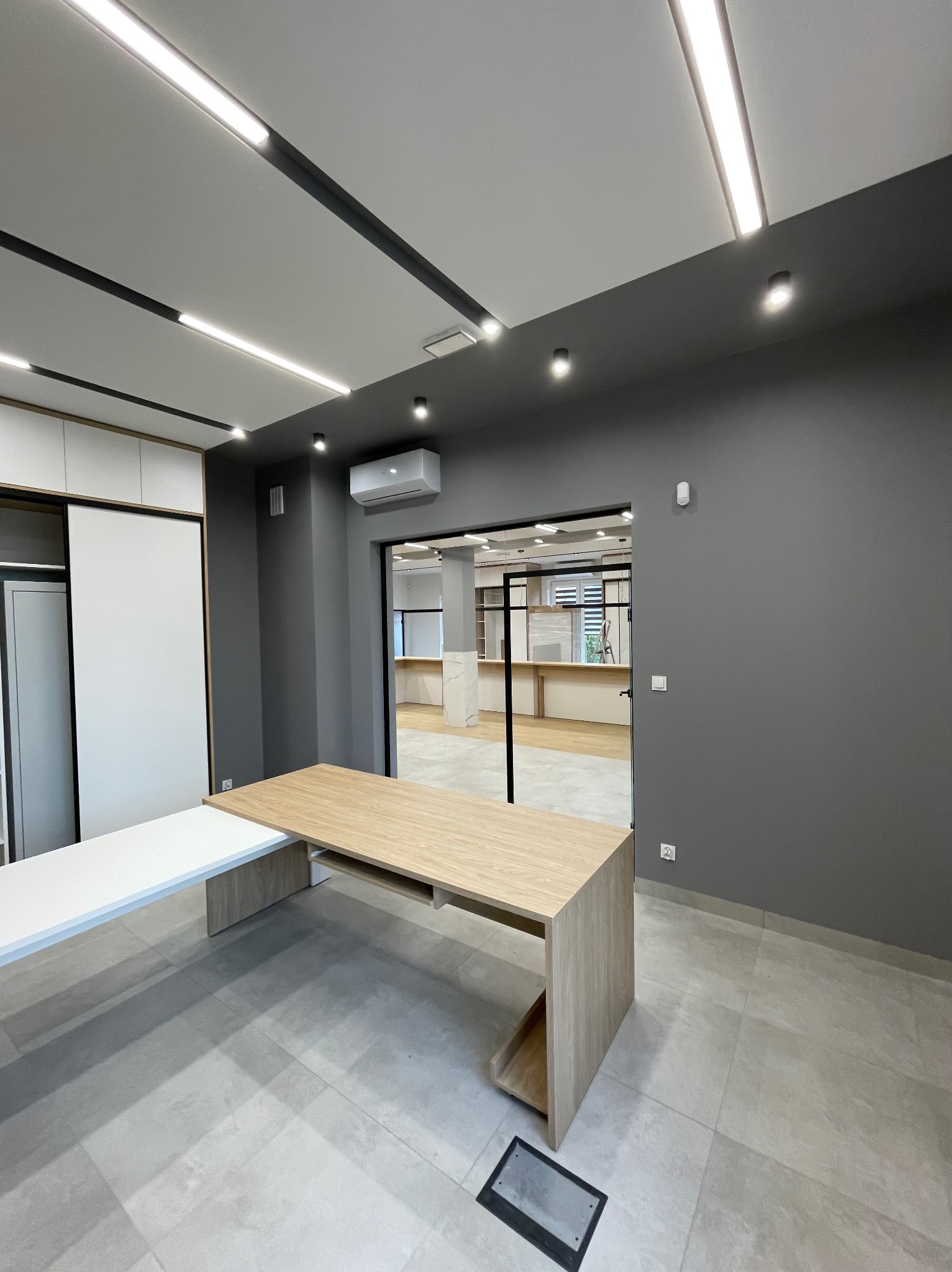 Nowoczesne biuro po remoncie: jasne, liniowe oświetlenie LED sufitowe, szare ściany, drewniane biurka. Widoczne gniazda elektryczne w podłodze i na ścianie.