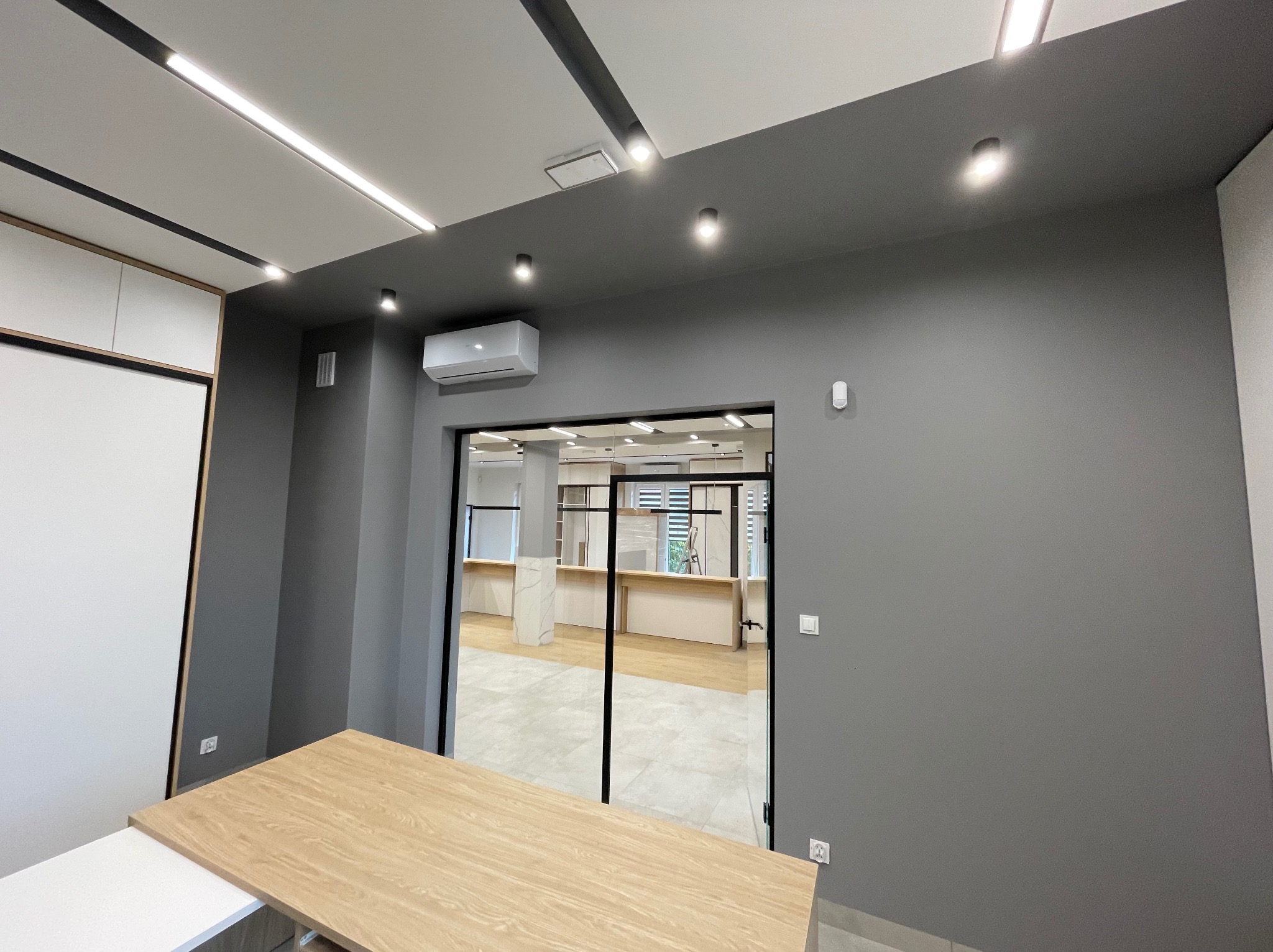 Nowoczesne biuro z oświetleniem LED:  liniowe lampy sufitowe i punktowe reflektory. Szara ściana, klimatyzator, gniazdka elektryczne i szklane drzwi do kolejnego pomieszczenia.