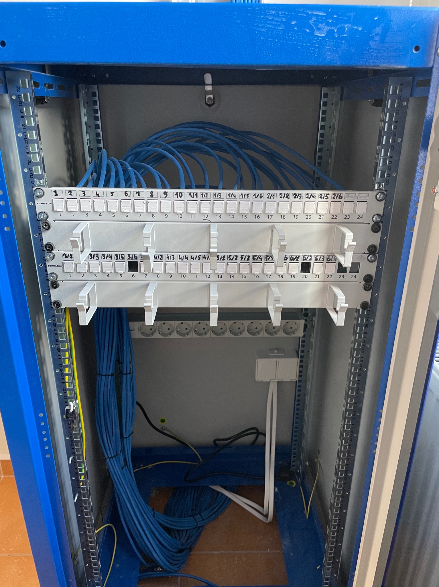 Panel krosowniczy w szafie rack z uporządkowanymi kablami Ethernet w kolorze niebieskim. Widoczne oznaczenia portów i moduły zasilające.