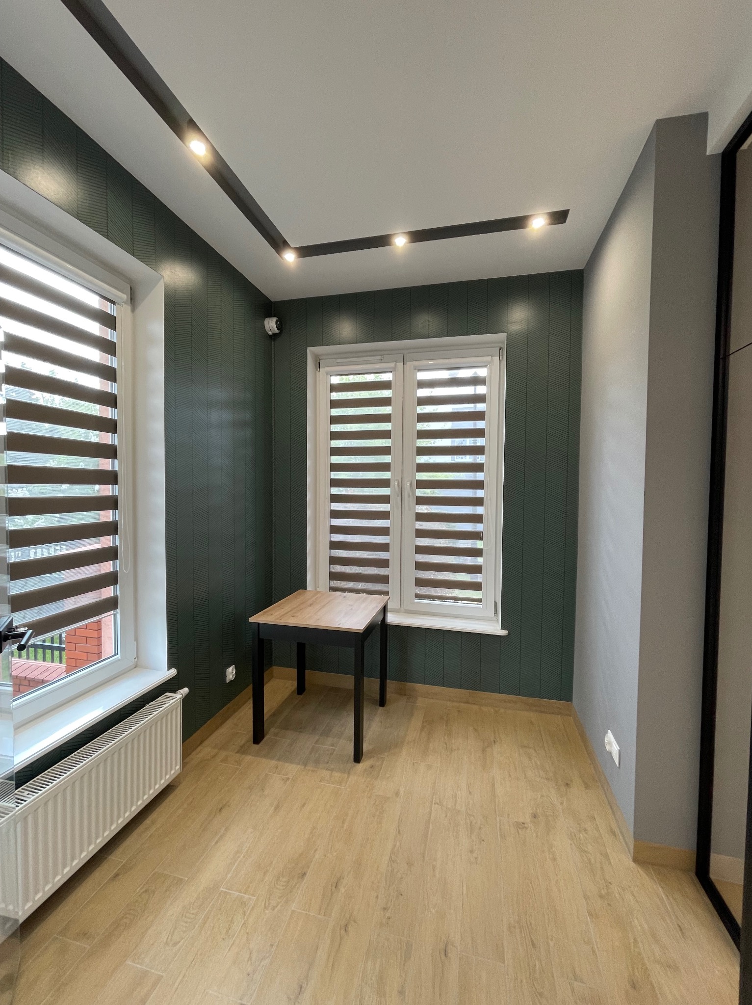 Nowoczesne wnętrze z oświetleniem punktowym w suficie, zieloną ścianą z wzorem, oknami z żaluzjami i małym stolikiem. Widoczny grzejnik i fragment instalacji elektrycznej.