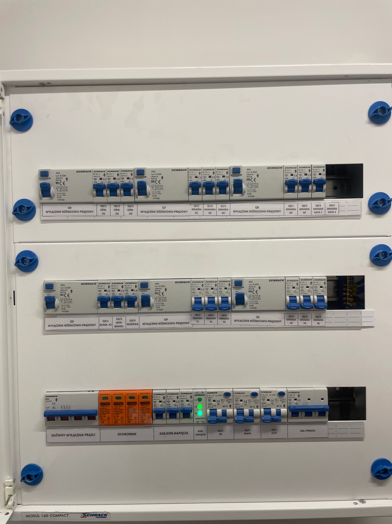 Rozdzielnica elektryczna Schrack z wyłącznikami różnicowoprądowymi i bezpiecznikami, z opisami obwodów, zamontowana w białej obudowie. Widoczne oznaczenia gniazd.