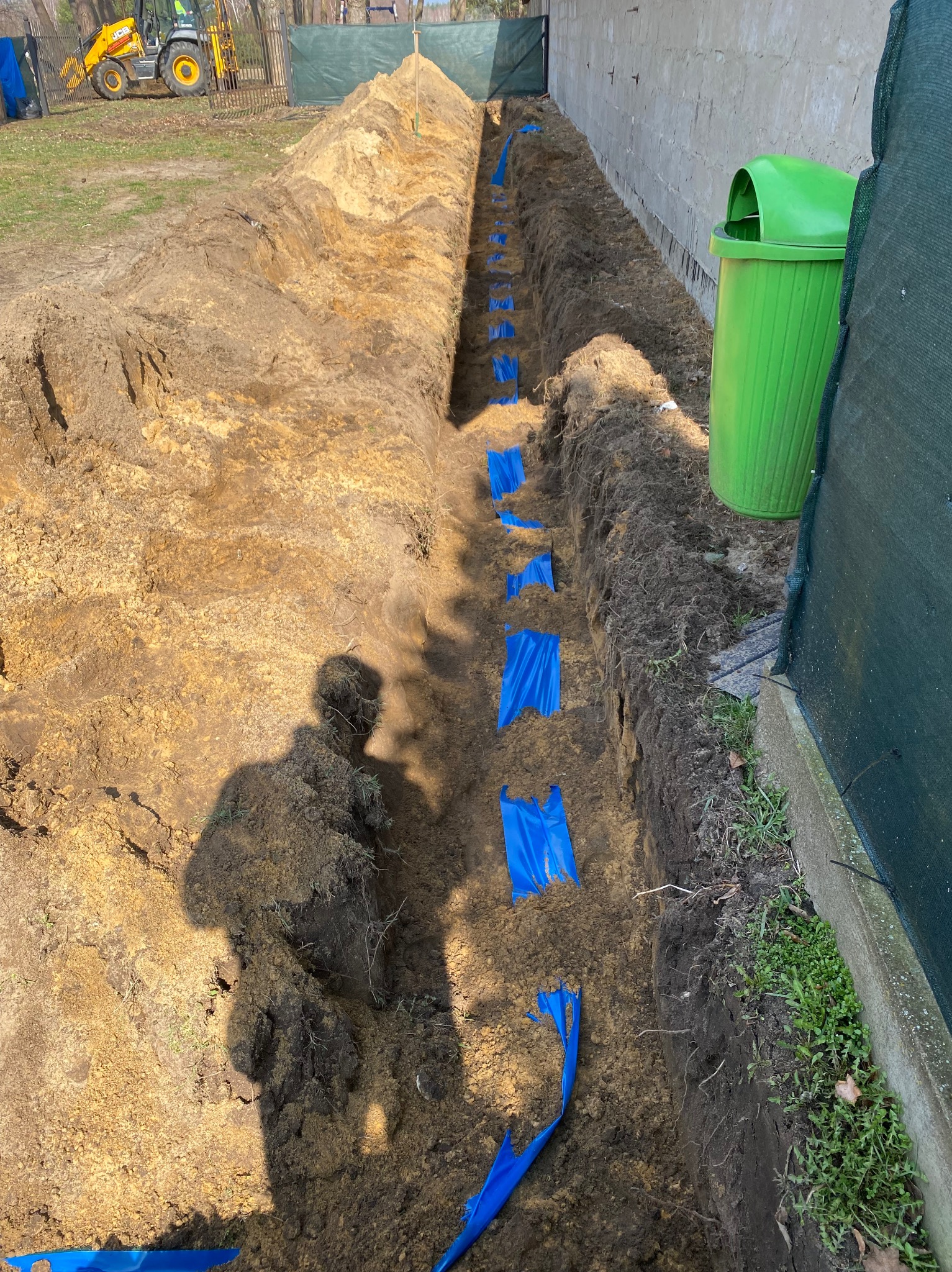 Wykop w ziemi z niebieskimi znacznikami, prawdopodobnie pod instalację. W tle widoczny sprzęt budowlany i zielony kosz na śmieci. Cień osoby w lewym dolnym rogu.