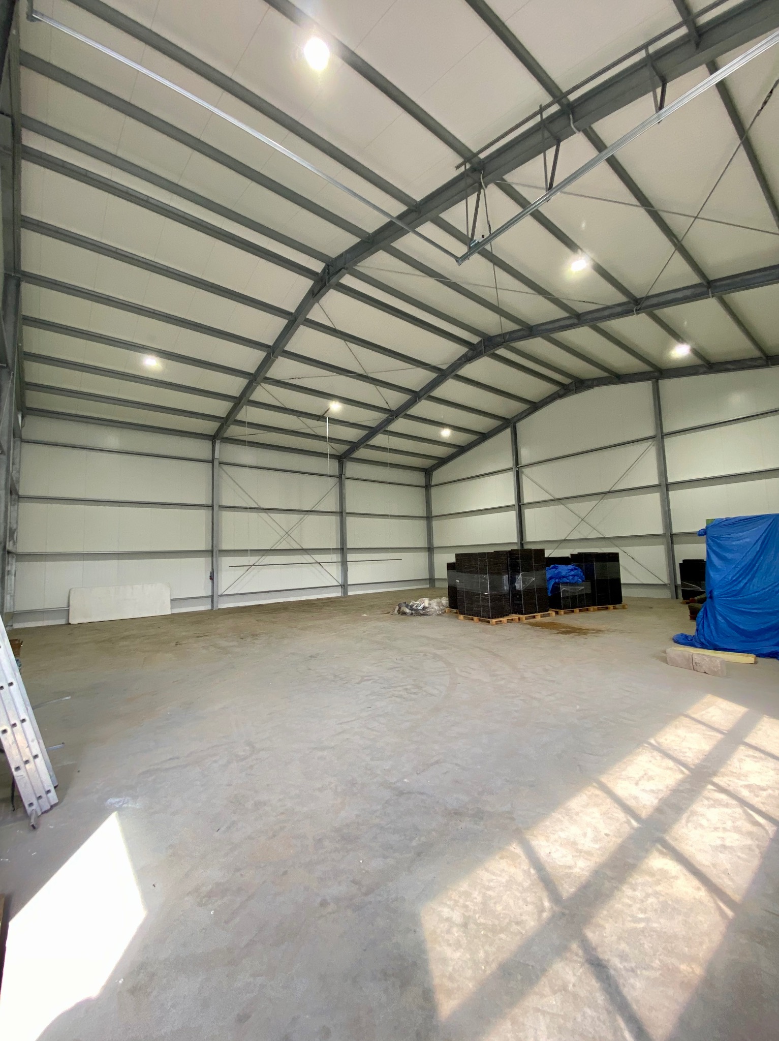 Wnętrze hali z metalową konstrukcją i oświetleniem LED. Widoczne materiały budowlane i drabina oparta o ścianę. Jasne, dzienne światło.