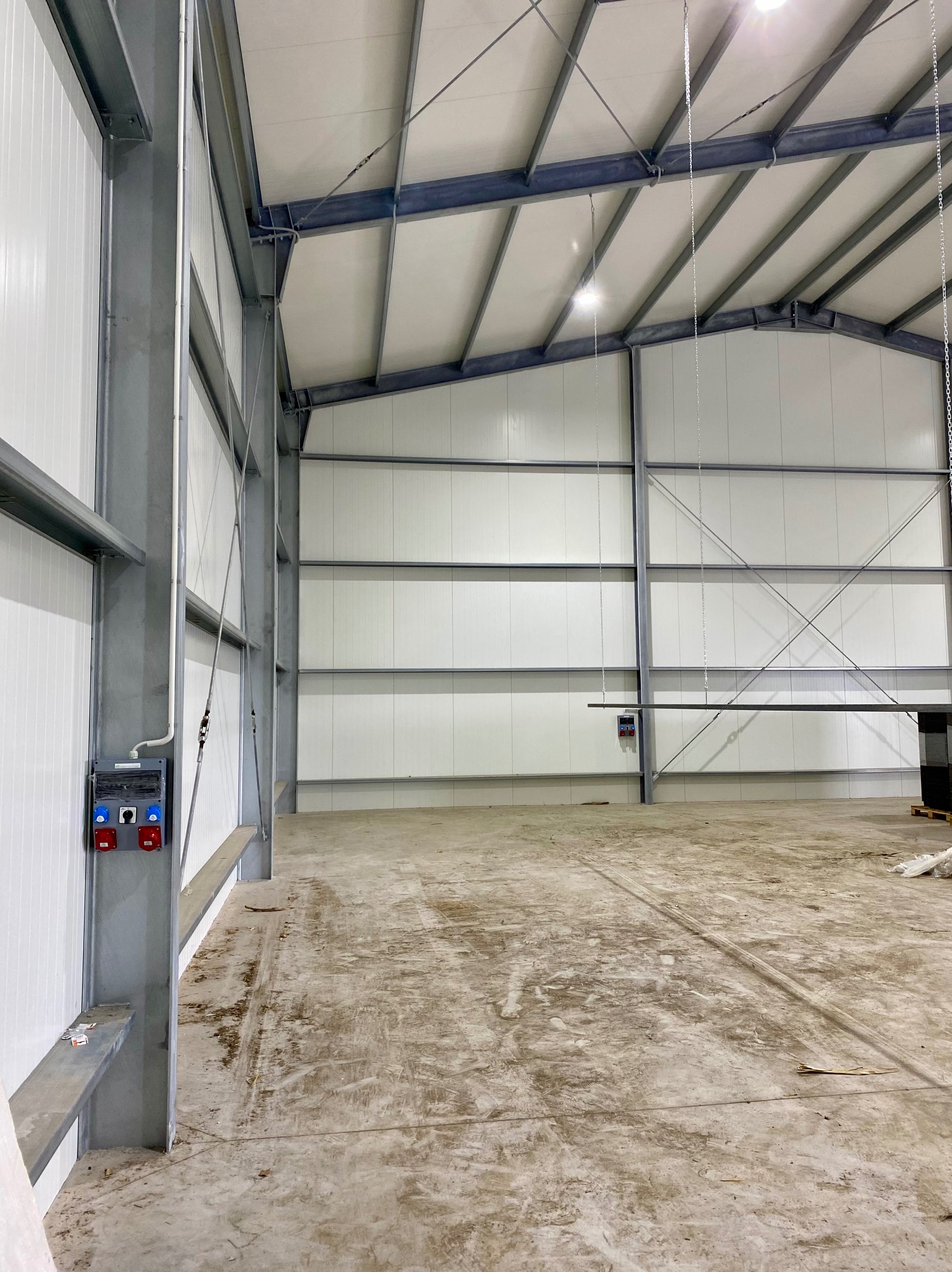 Wnętrze hali produkcyjnej z widoczną instalacją elektryczną: szara skrzynka z czerwonymi i niebieskimi gniazdami na słupie, białe ściany z paneli, metalowa konstrukcja dachu.
