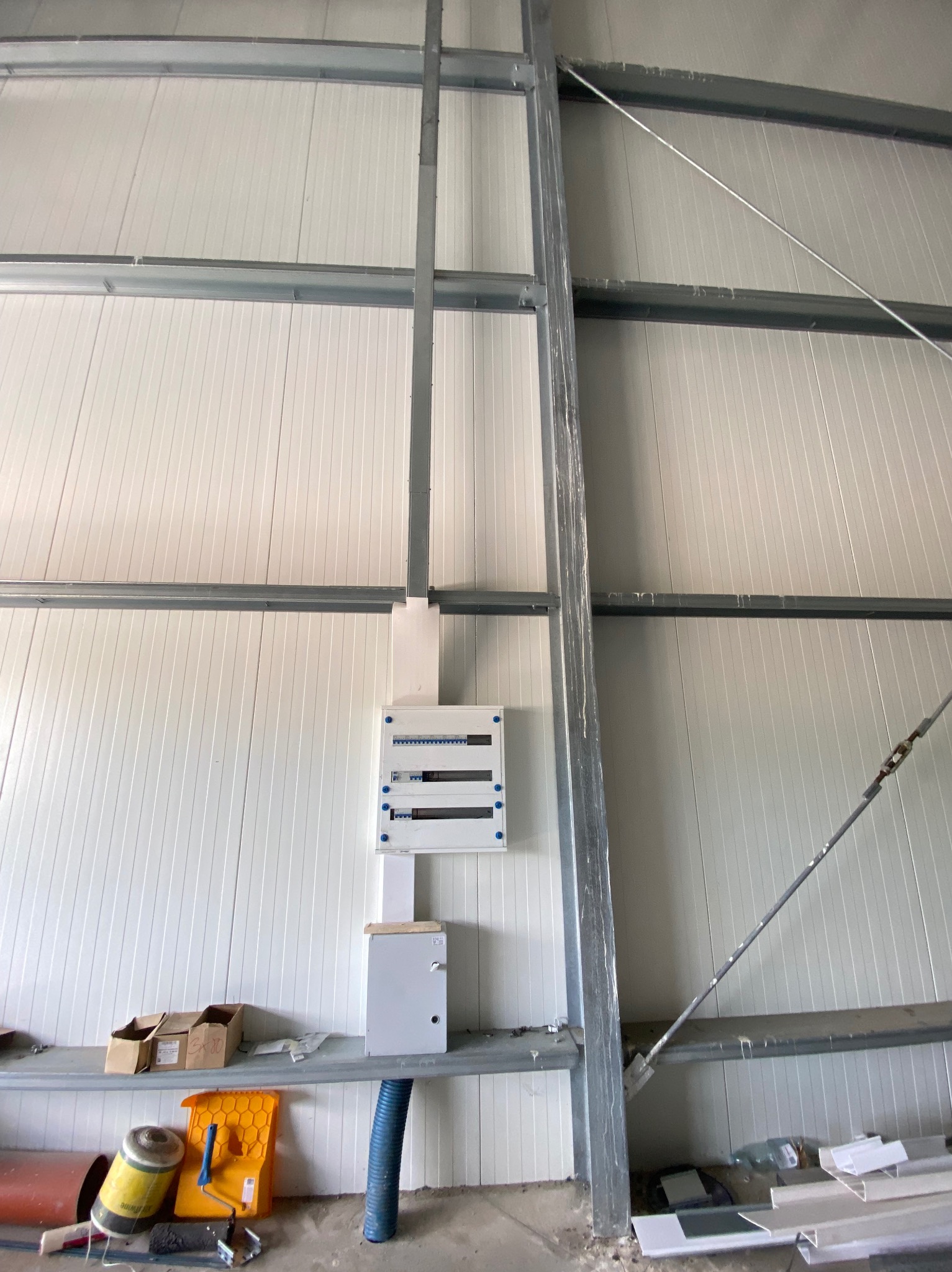 Rozdzielnia elektryczna zamontowana na ścianie hali z blachy trapezowej, widoczne kable w peszlu i narzędzia na półce. Instalacja w trakcie realizacji.