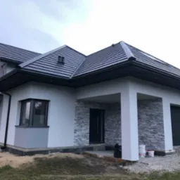 Nowoczesny dom jednorodzinny z szarą elewacją, ciemną dachówką i elementami dekoracyjnymi z kamienia, widoczne puszki farb na podjeździe.