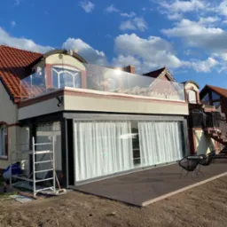 Nowoczesny taras na dachu budynku z przeszkloną balustradą, drewnianą podłogą i czarnymi krzesłami, widok na błękitne niebo z chmurami, w tle sąsiednie budynki, na pierwszym planie rozkopana ziemia...