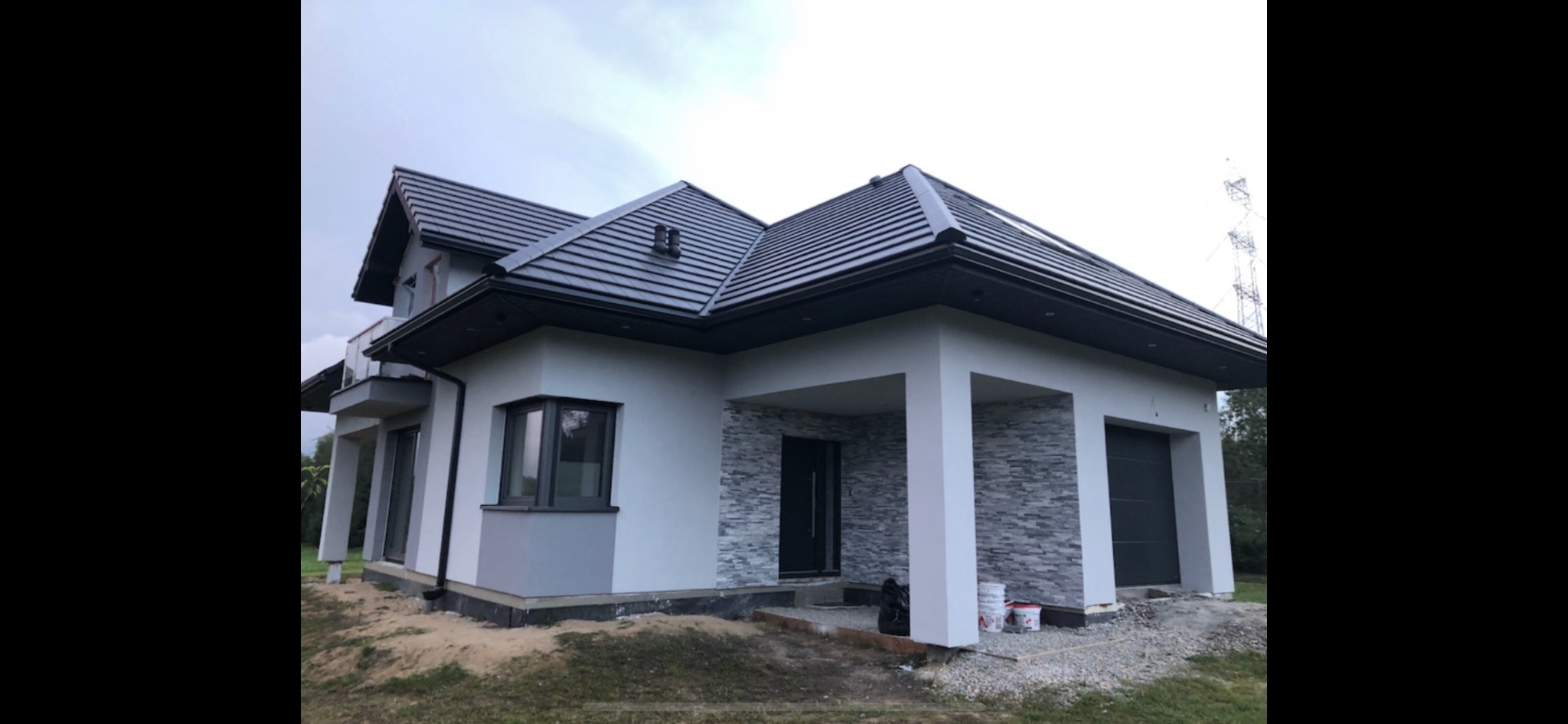 Nowoczesny dom jednorodzinny z szarą elewacją, ciemną dachówką i elementami dekoracyjnymi z kamienia, widoczne puszki farb na podjeździe.