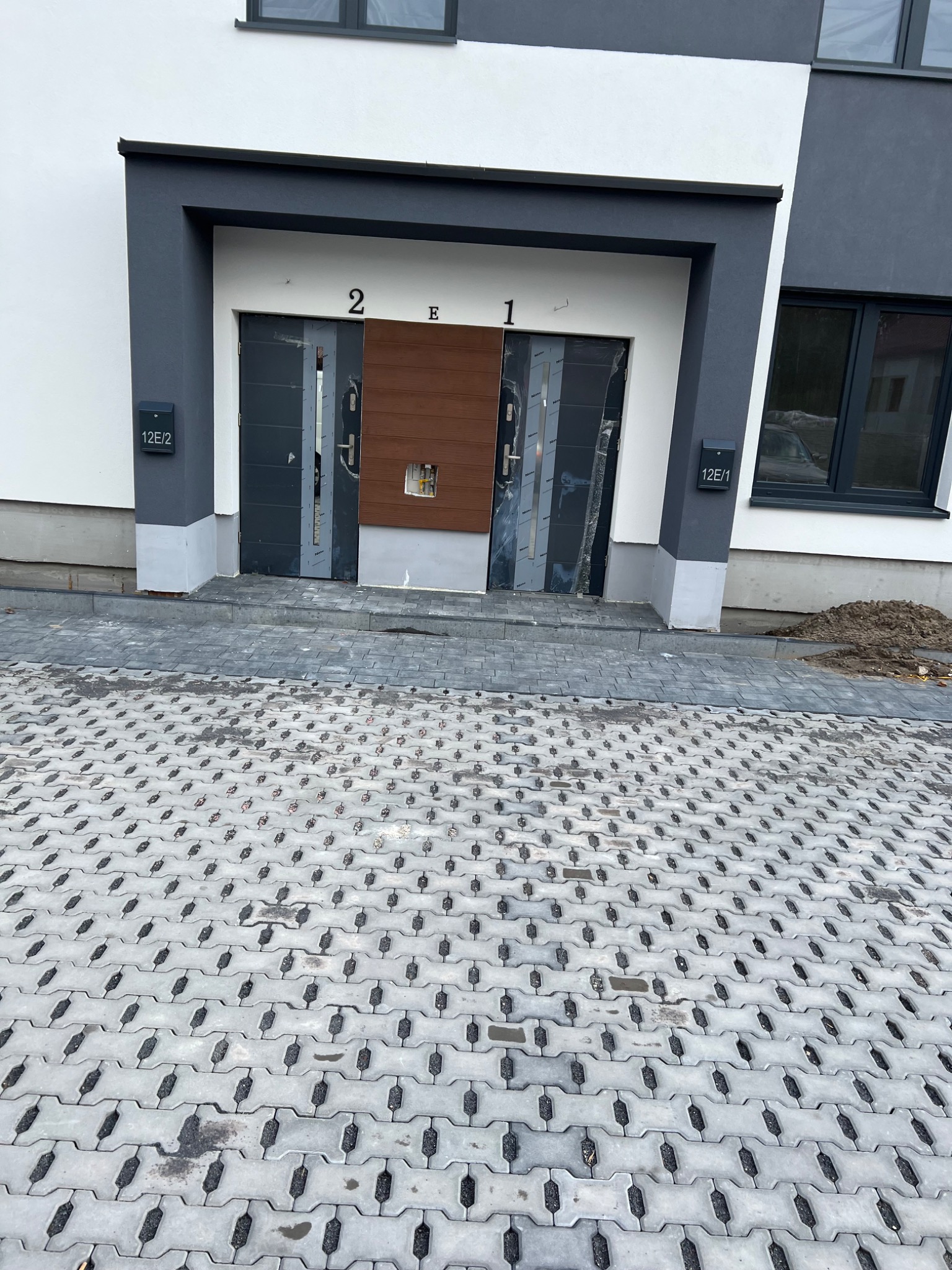 Wejście do budynku z otwartymi, szarymi drzwiami, drewnianym panelem i numerami 2E1 nad wejściem, widoczne skrzynki pocztowe 12E/1 i 12E/2, szara kostka brukowa na podjeździe.