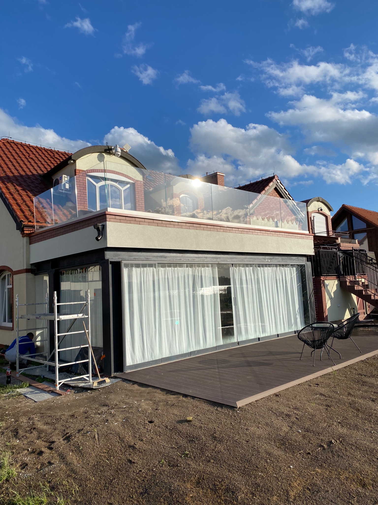 Nowoczesny taras na dachu budynku z przeszkloną balustradą, drewnianą podłogą i czarnymi krzesłami, widok na błękitne niebo z chmurami, w tle sąsiednie budynki, na pierwszym planie rozkopana ziemia...