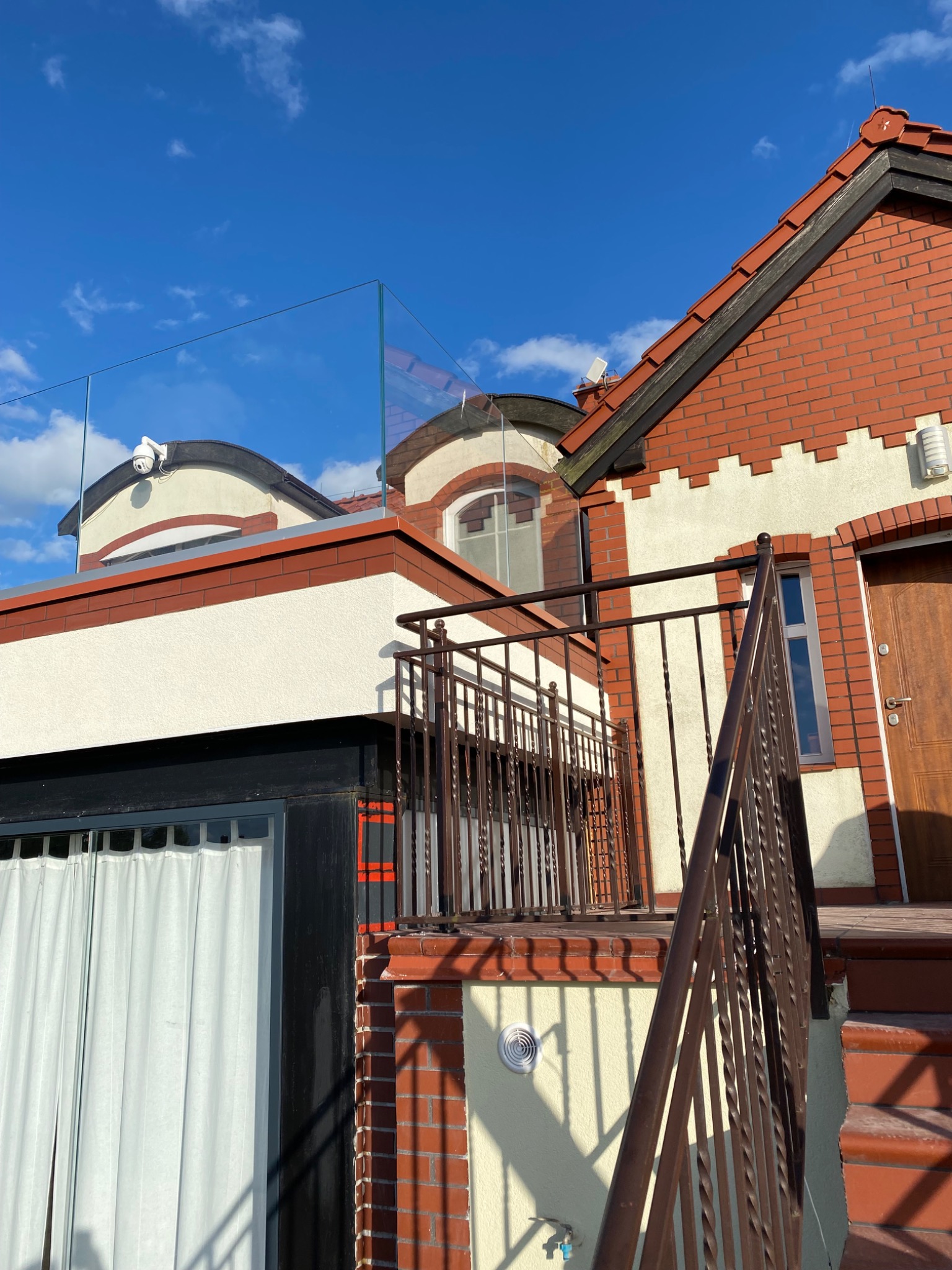 Dwupiętrowy budynek z czerwonej cegły i białego tynku, z balkonem z metalową balustradą i szklaną barierką na dachu, widok z dołu w słoneczny dzień.