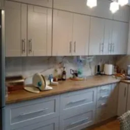 Szara zabudowa kuchenna z drewnianym blatem, widoczne naczynia na blacie i nowoczesna lampa sufitowa.