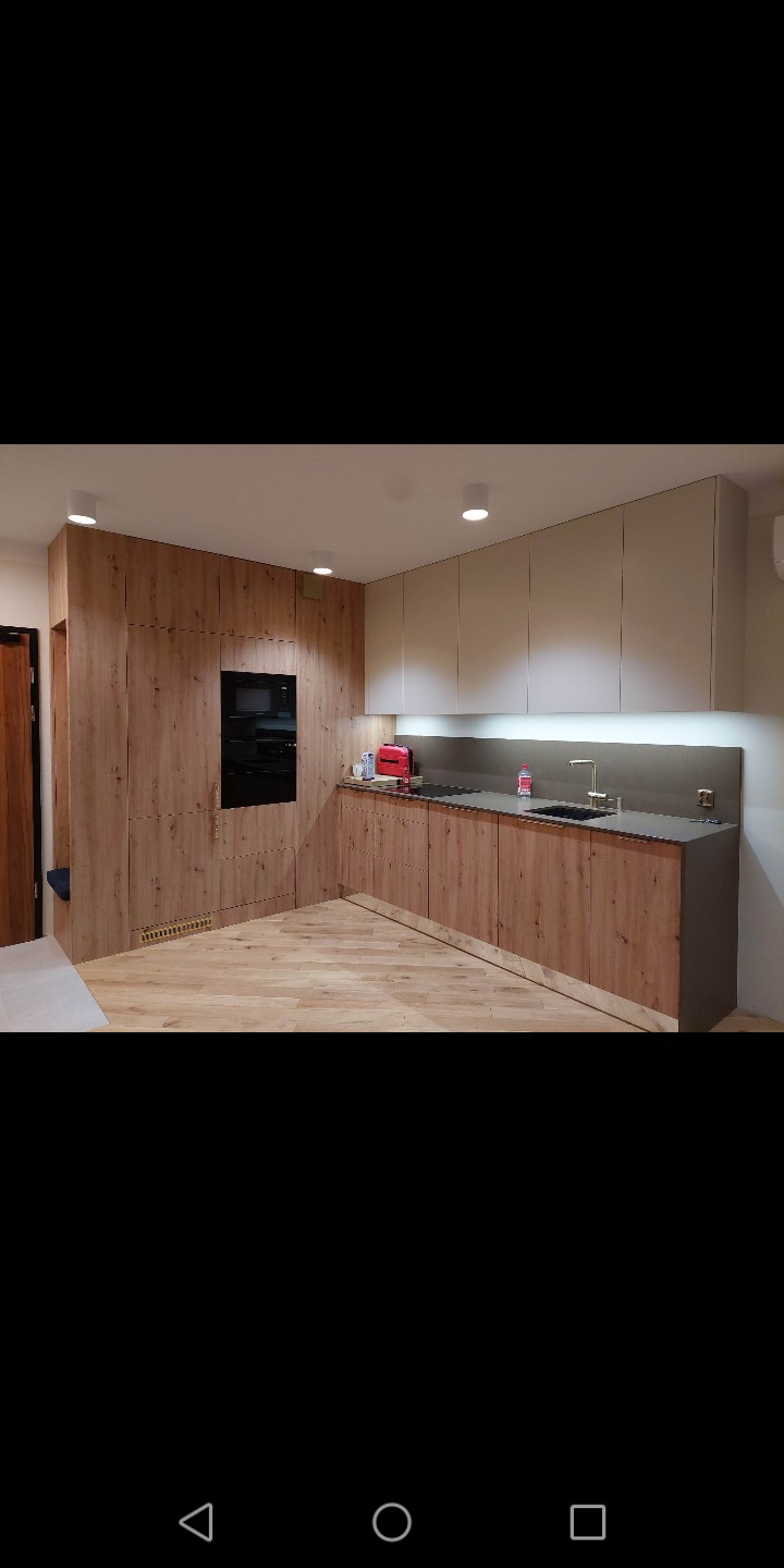 Nowoczesna kuchnia z frontami szafek w kolorze jasnego drewna z czarnym piekarnikiem w zabudowie i białymi szafkami wiszącymi, oświetlona punktowymi lampami.