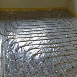 Białe rury ogrzewania podłogowego ułożone na srebrnej folii z czerwoną siatką, zabezpieczone żółtą taśmą przy ścianach w surowym wnętrzu z oknem.