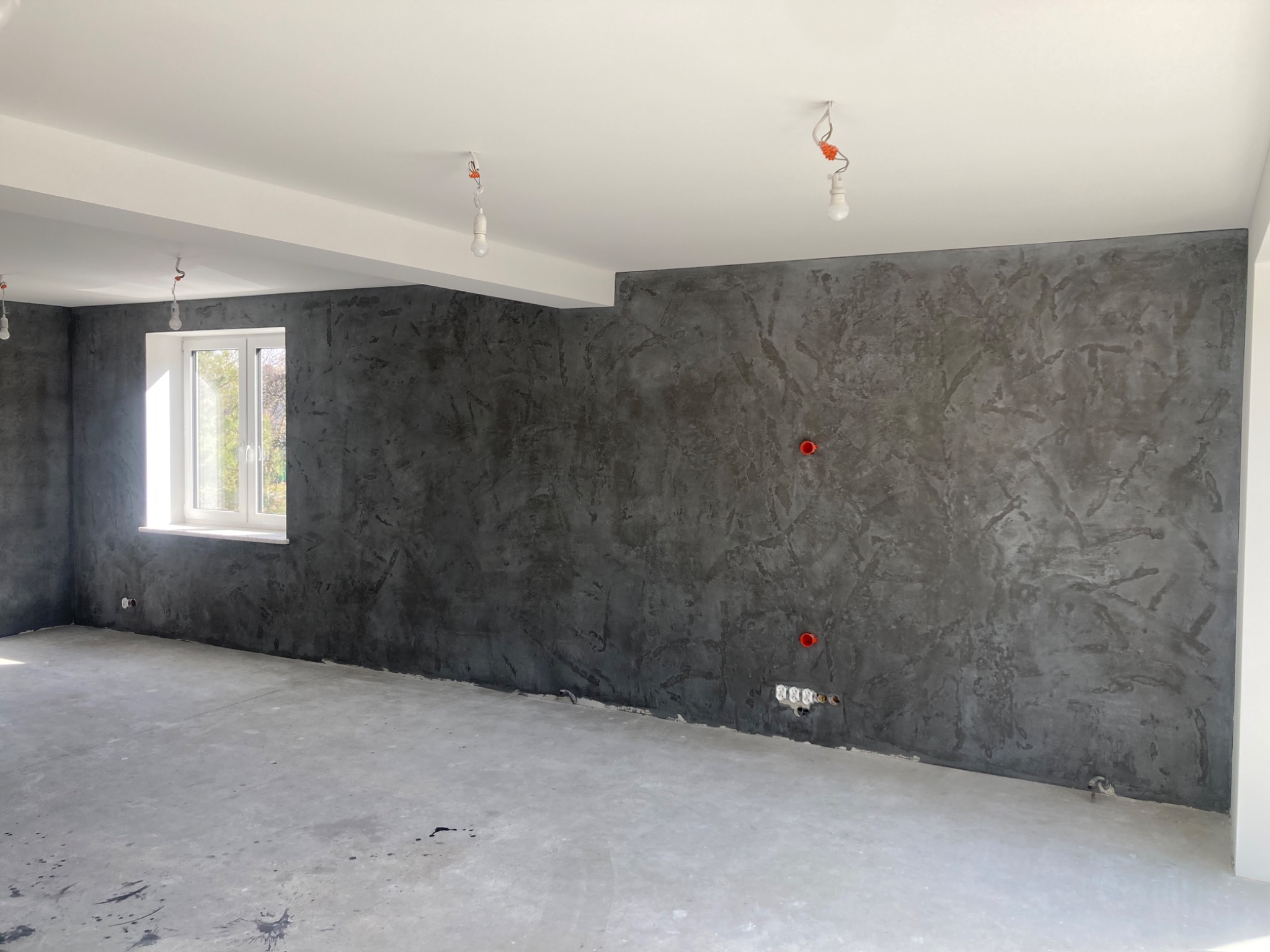 Surowe wykończenie ścian w odcieniach szarości z widocznymi instalacjami elektrycznymi, betonowa podłoga w stanie surowym, białe okno i sufit z wiszącymi żarówkami.
