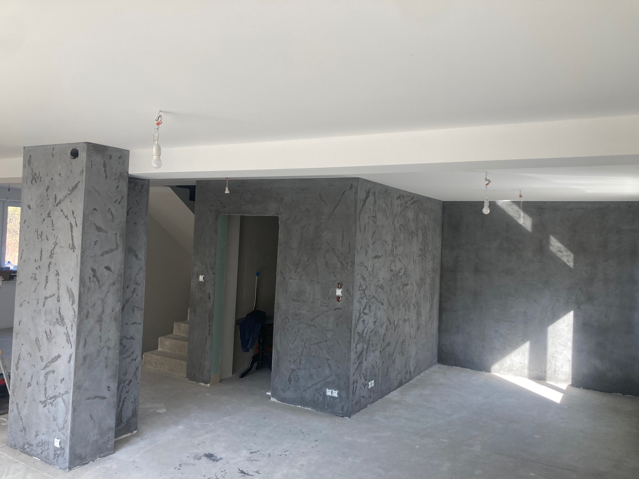Surowe wnętrze w trakcie wykańczania: szare ściany z fakturą, surowy beton na podłodze, wiszące żarówki bez kloszy, widoczne schody i otwór drzwiowy.