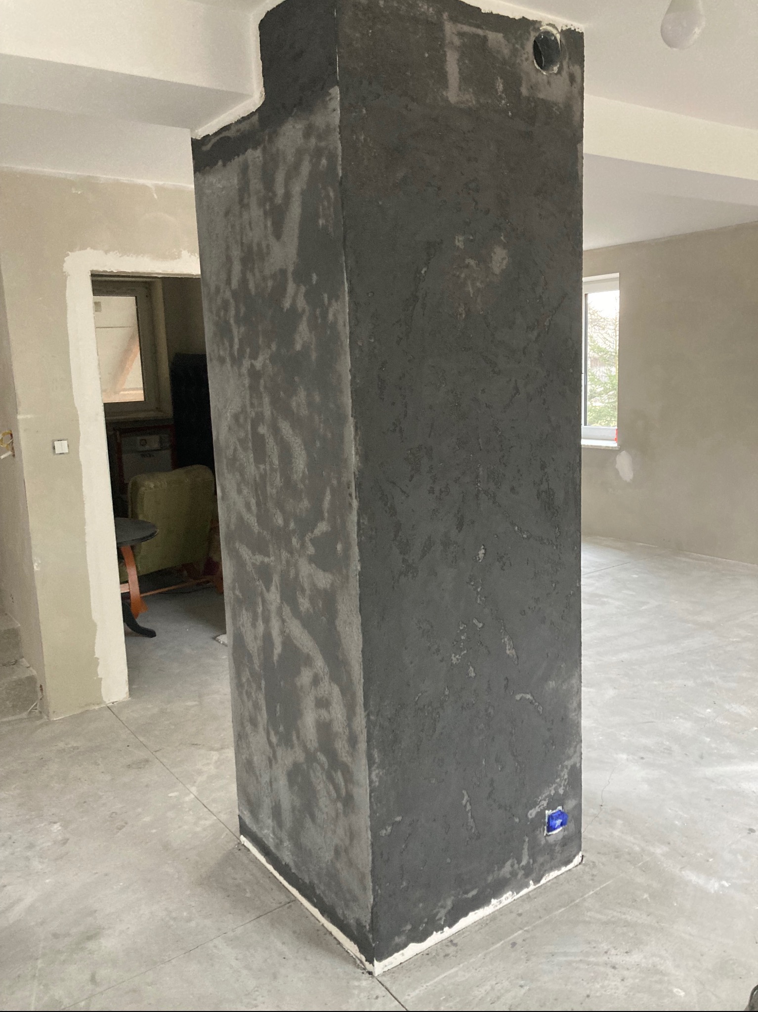 Surowy, ciemnoszary filar w trakcie wykończenia, widoczny otwór instalacyjny w górnej części, wnętrze w stanie deweloperskim z surowymi ścianami i podłogą.