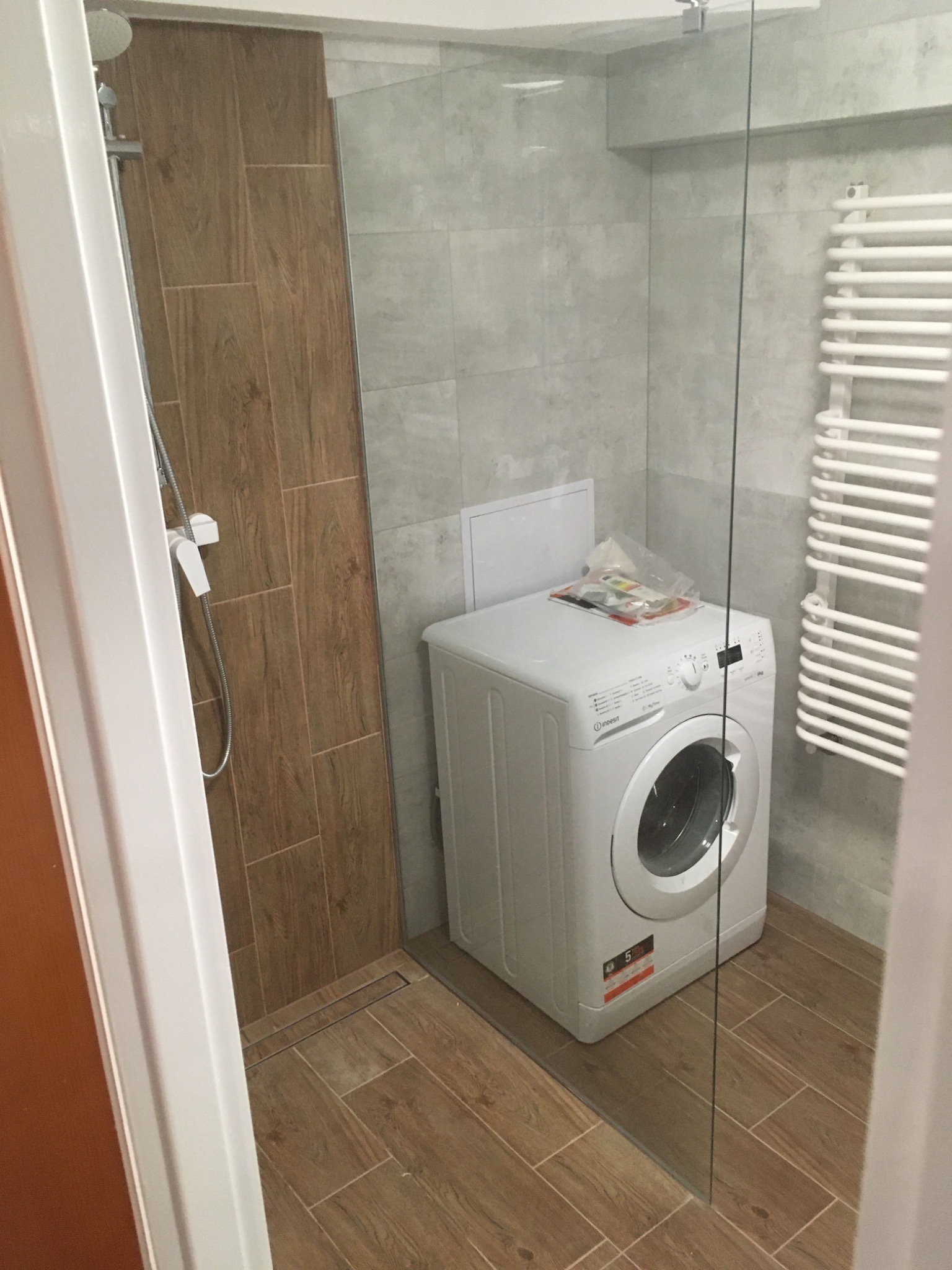 Wnętrze łazienki z kabiną prysznicową, grzejnikiem drabinkowym i pralką Indesit, wykończone płytkami imitującymi drewno i szarym gresem.