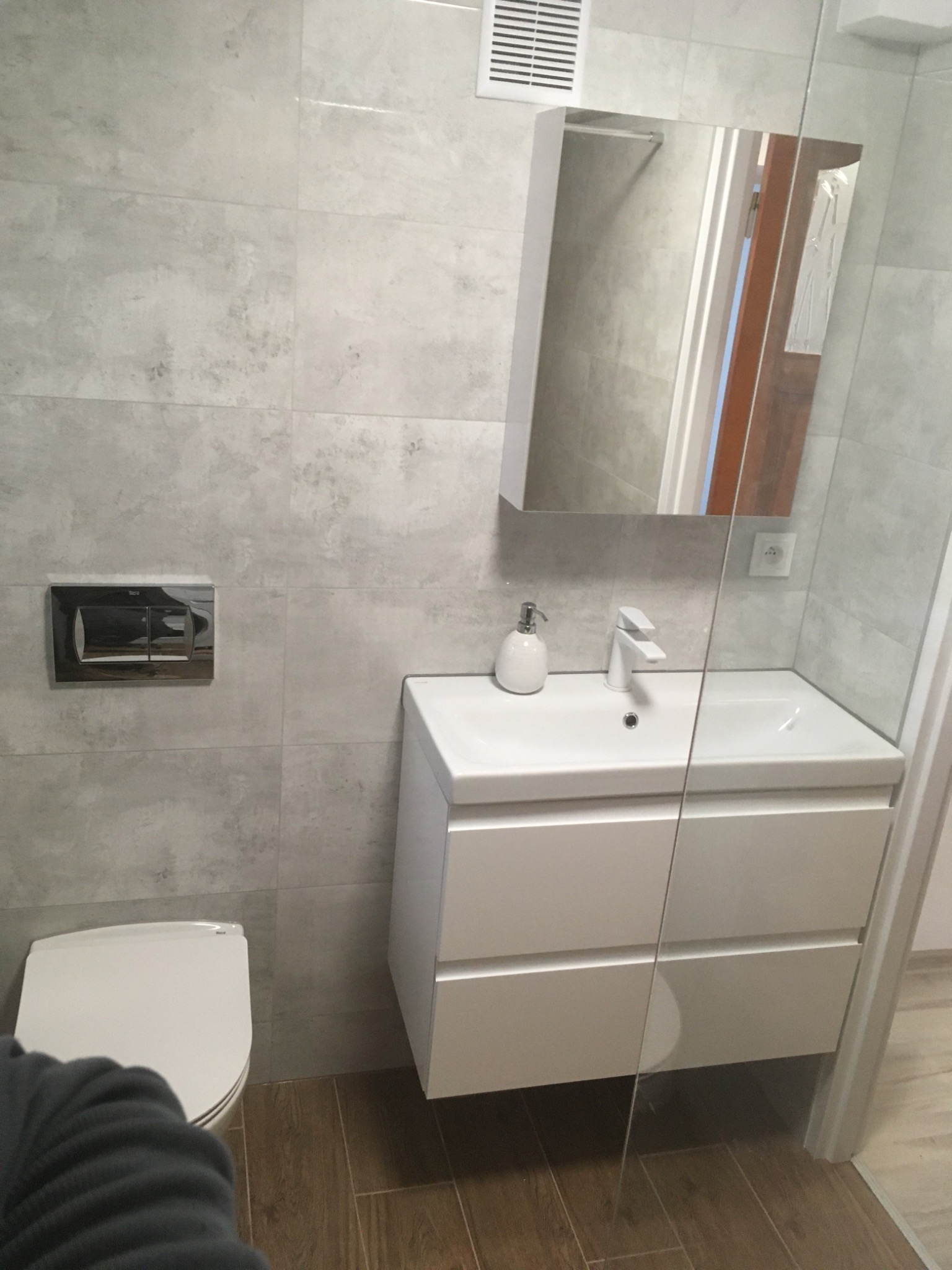 Nowoczesna łazienka z szarymi płytkami imitującymi kamień, białą szafką pod umywalką, wiszącą toaletą i lustrem z oświetleniem wbudowanym w szafkę.