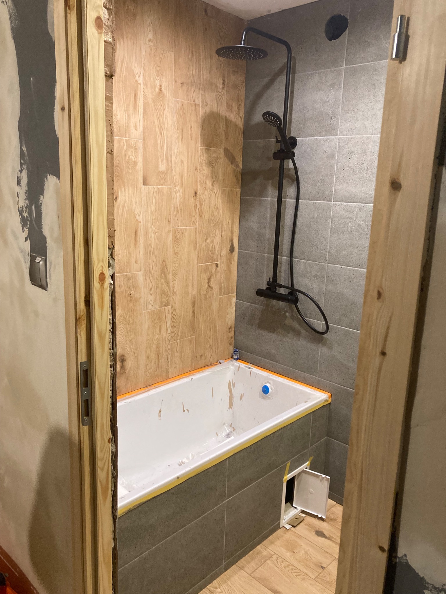 Remont łazienki w trakcie: biała wanna z pomarańczowym zabezpieczeniem, czarna bateria prysznicowa na szarych płytkach, drewnopodobne płytki na ścianie, otwarta futryna drzwiowa, widoczny otwór...