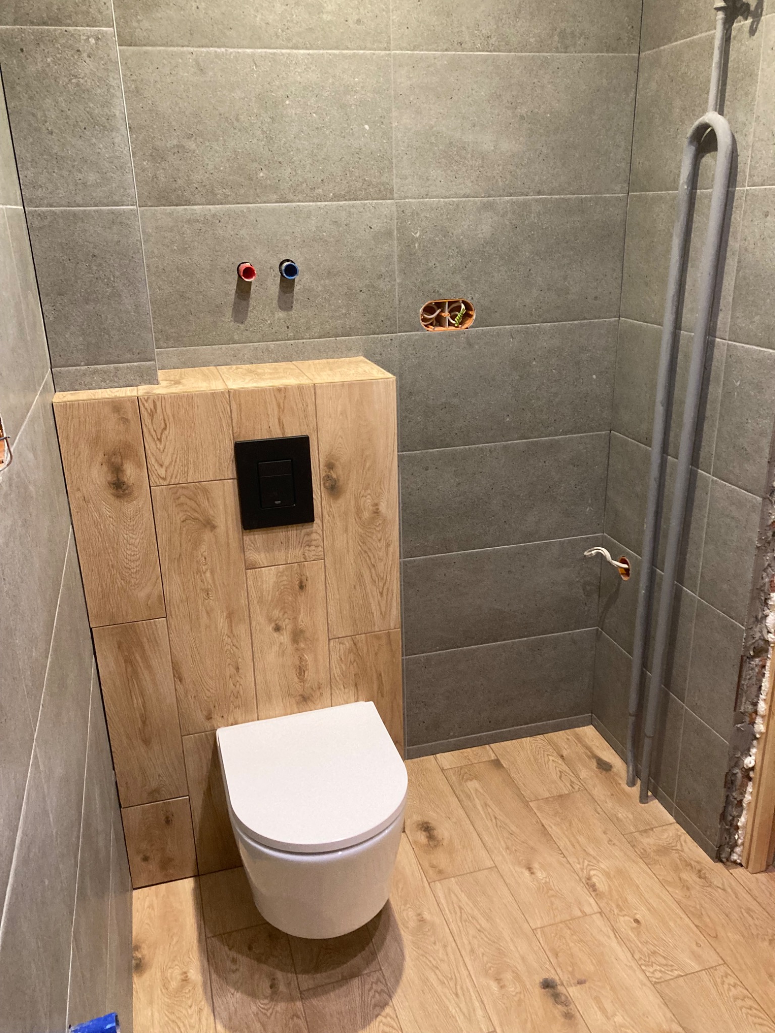 Podtynkowa toaleta wisząca z czarnym przyciskiem spłukującym, obudowana płytami imitującymi drewno, na tle szarych płytek ściennych i drewnopodobnej podłogi. Widoczne instalacje hydrauliczne...