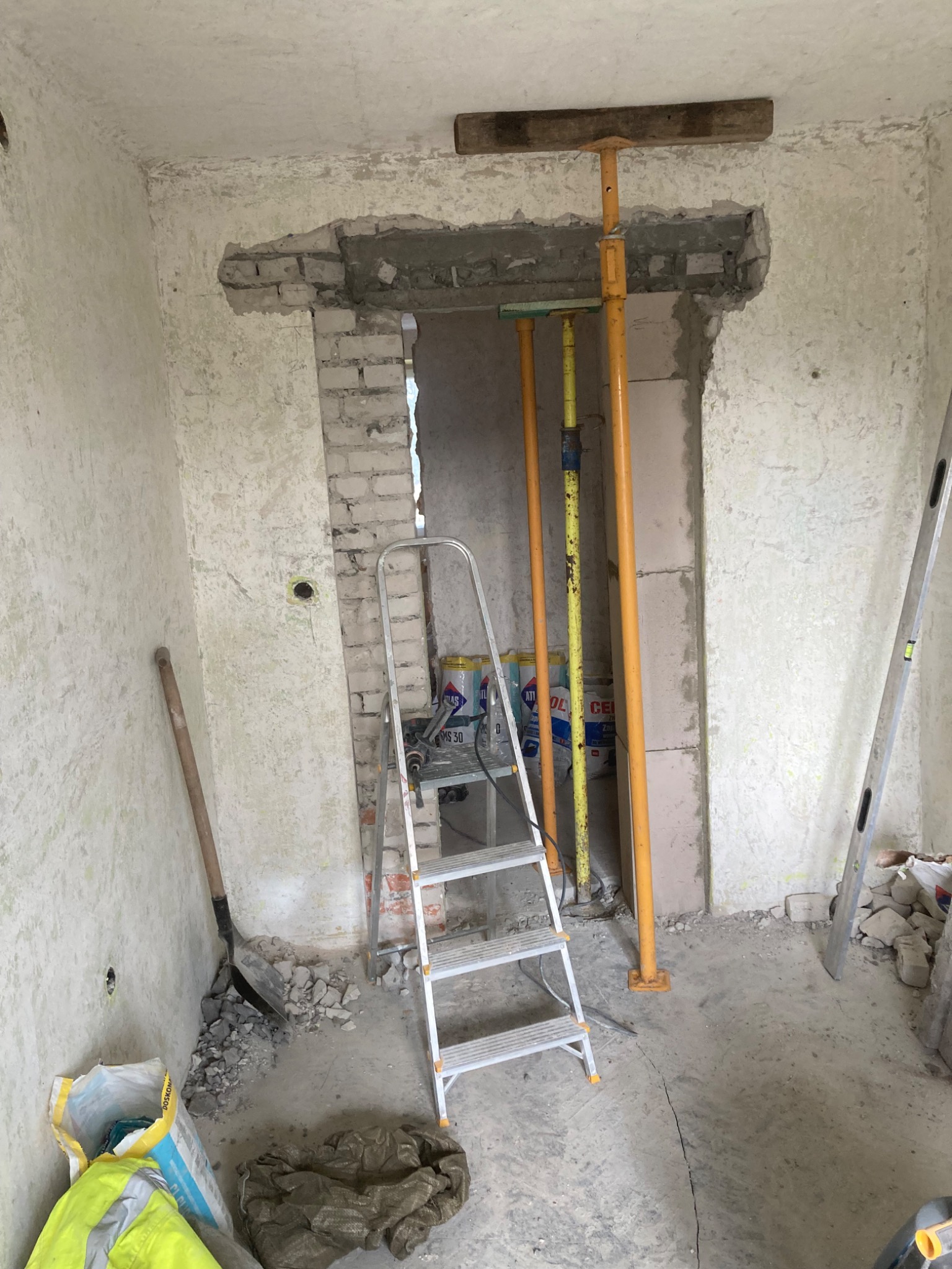 Wnętrze w trakcie prac murarskich: wybite przejście w ścianie podparte stemplami budowlanymi, drabina aluminiowa, gruz i narzędzia murarskie na podłodze, ściany z widoczną strukturą.