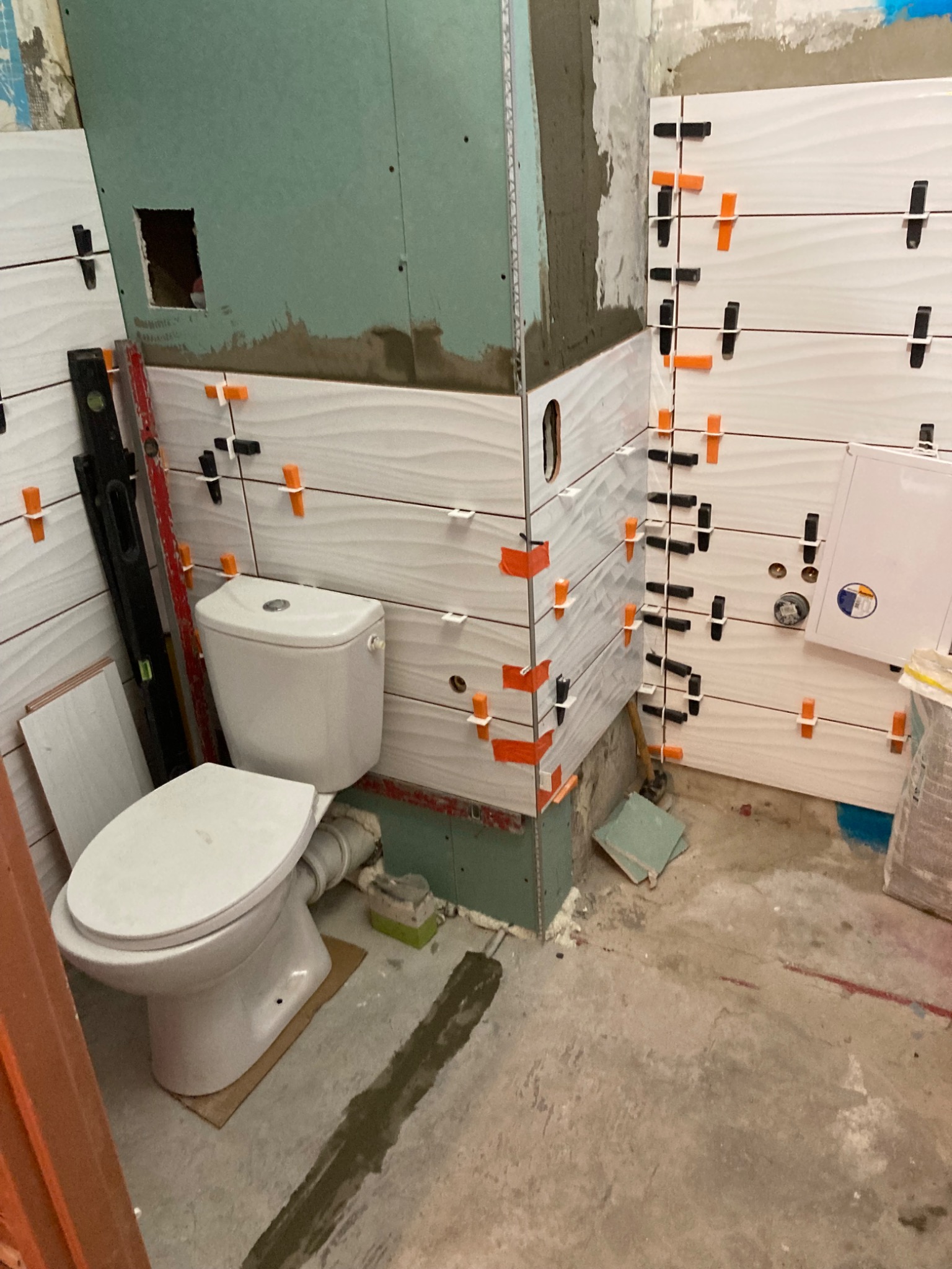 Montaż białych, falistych płytek w łazience, widoczne poziomice i krzyżyki dystansowe, częściowo zamontowana toaleta, surowy beton na podłodze.