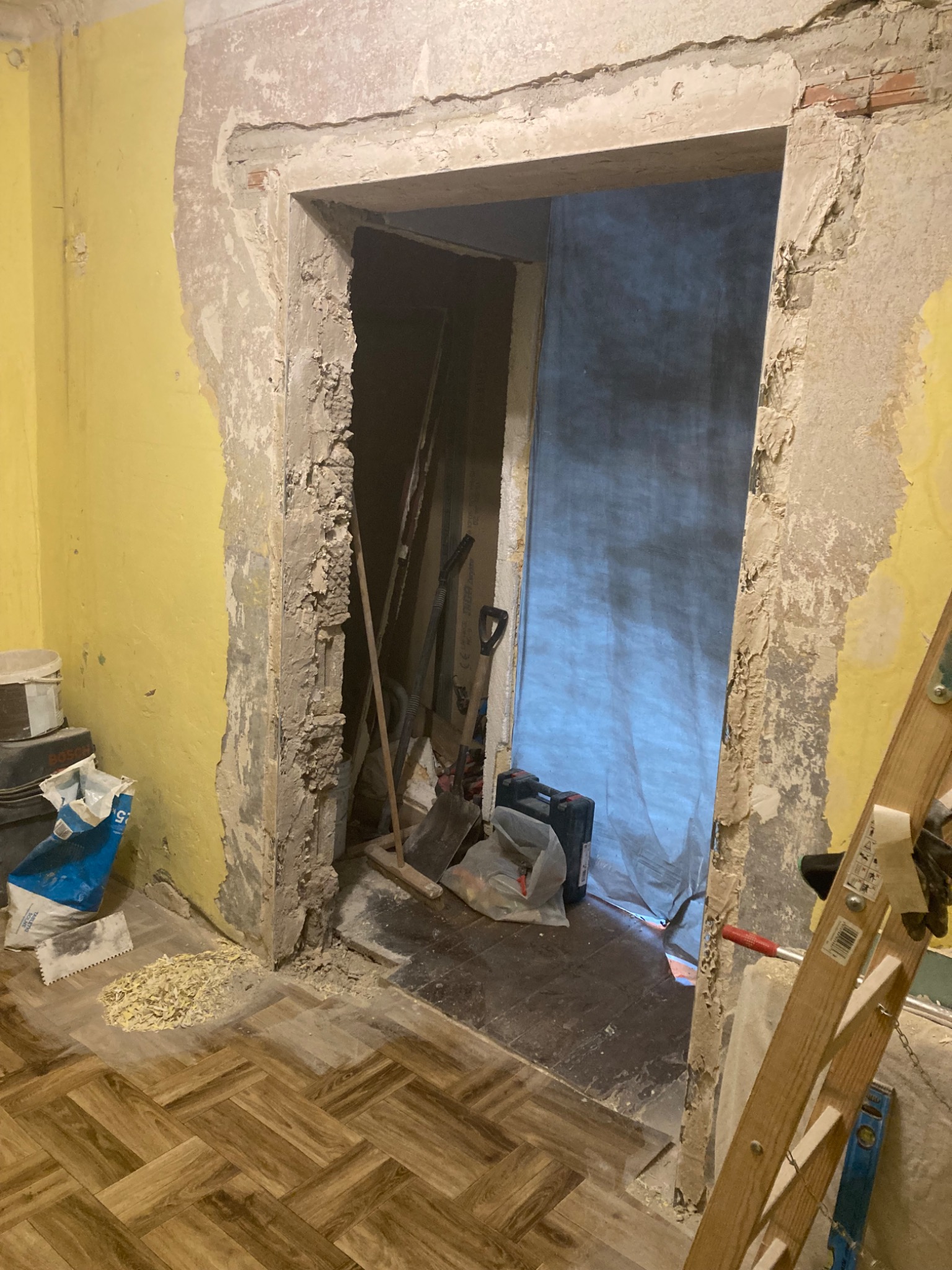 Demontaż ościeżnicy w trakcie remontu, widoczne resztki zaprawy, żółta ściana, parkiet w jodełkę, drabina oparta o ścianę i narzędzia w przejściu.