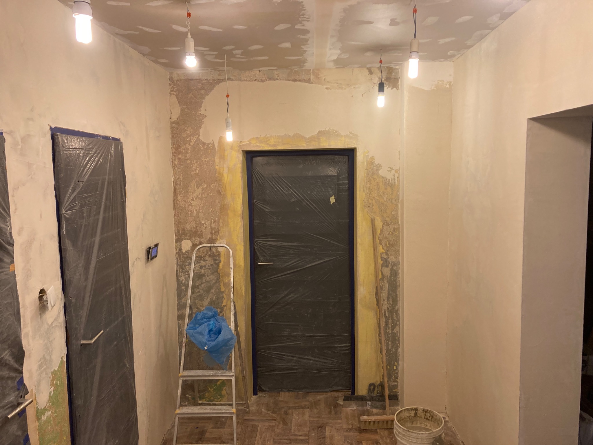 Pokój w trakcie szpachlowania ścian i sufitu, zabezpieczone drzwi folią, drabina z workiem, oświetlenie żarówkami na kablach.