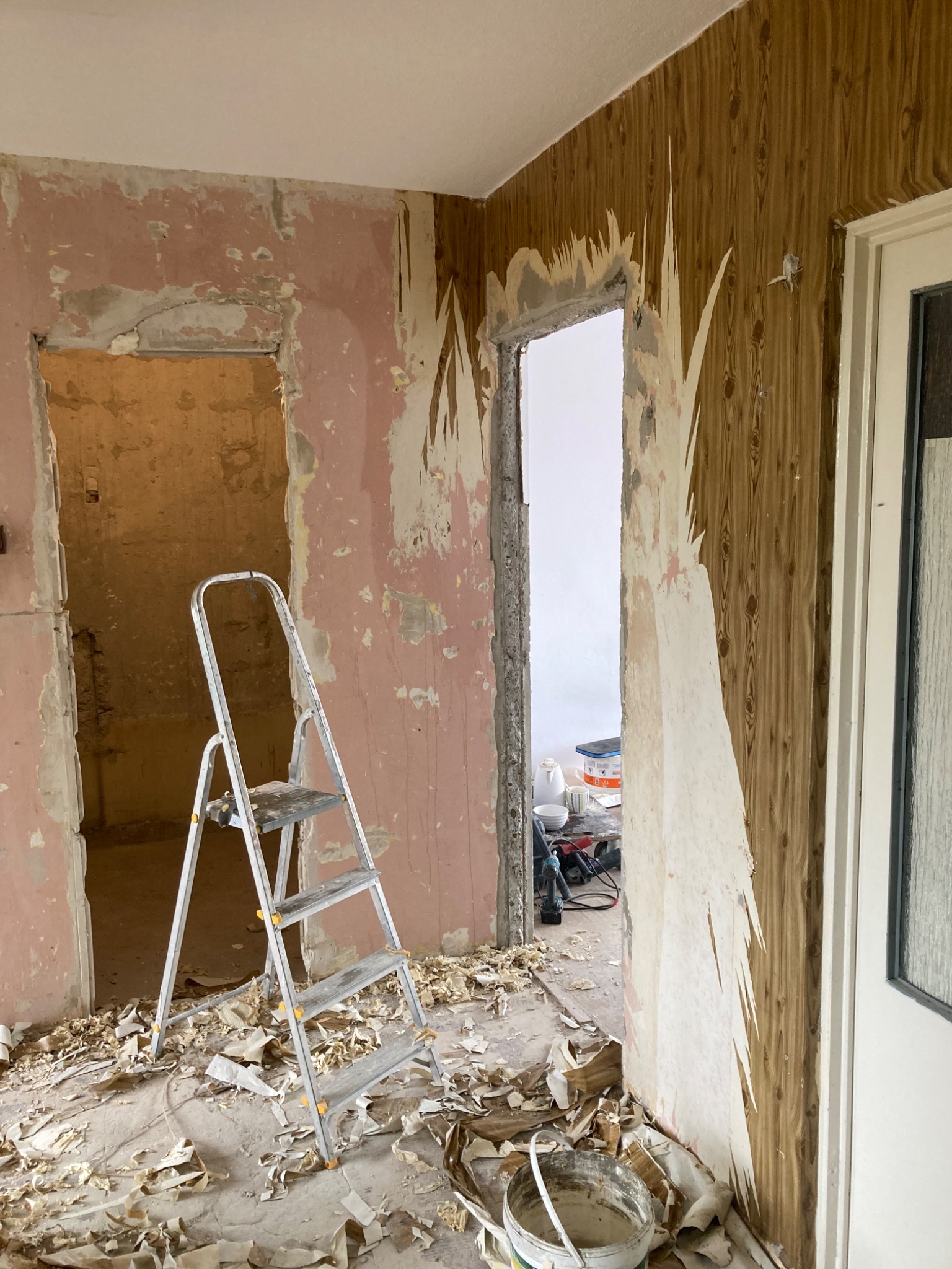 Pokój w trakcie remontu: zerwane tapety, różowe ściany, aluminiowa drabina, narzędzia w tle.