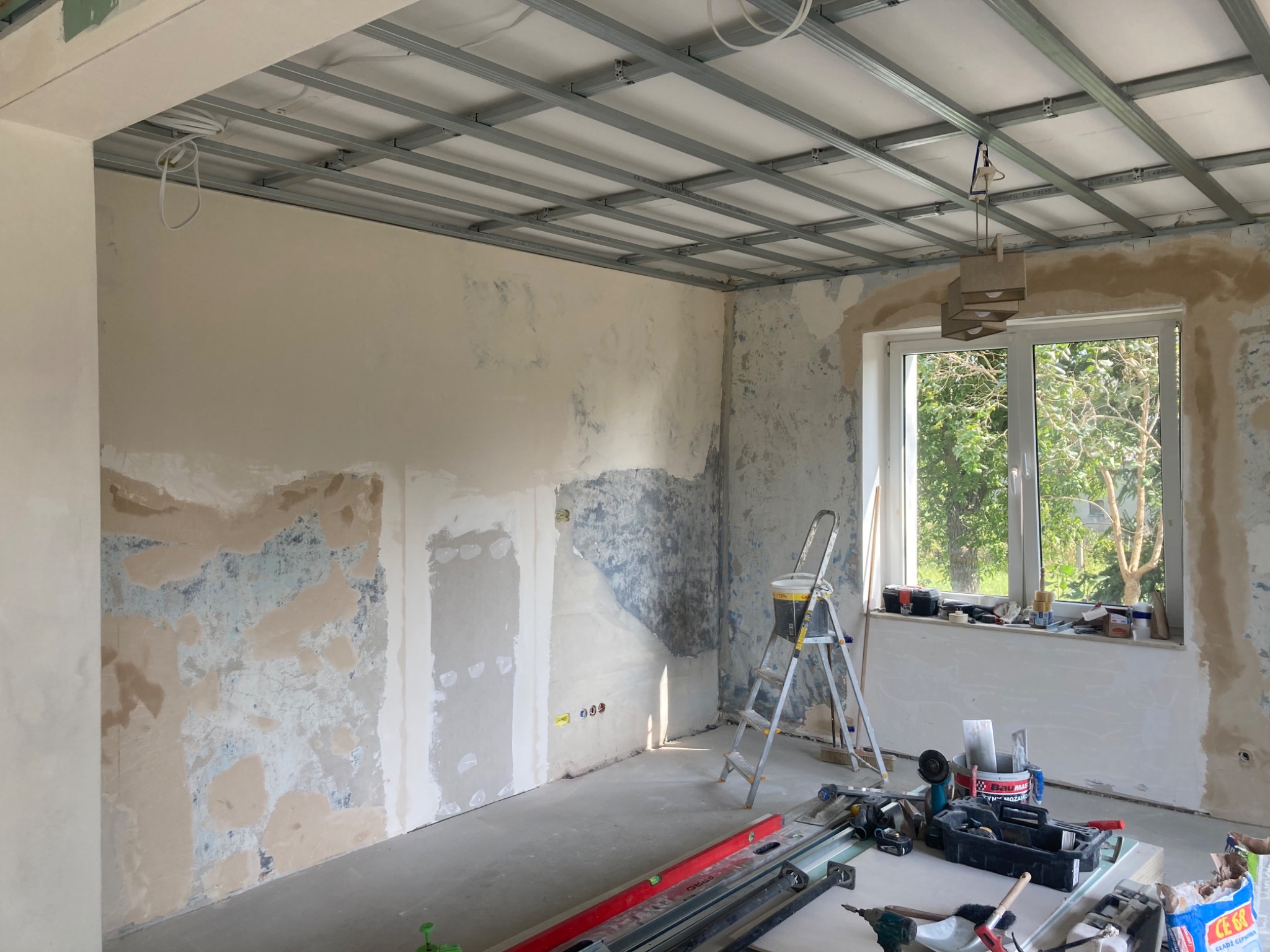 Pokój w trakcie przygotowania do malowania: częściowo otynkowane ściany, metalowy stelaż sufitu podwieszanego, drabina, narzędzia i materiały budowlane na podłodze.