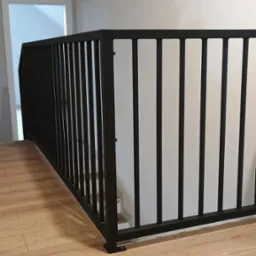 Nowoczesna, czarna balustrada schodowa z pionowych prętów, zamontowana w jasnym wnętrzu z panelami podłogowymi i widocznym fragmentem schodów.