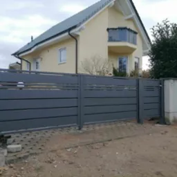 Nowoczesna, szara brama wjazdowa z poziomych paneli, widoczna na tle jasnego domu z balkonem i kostki brukowej.