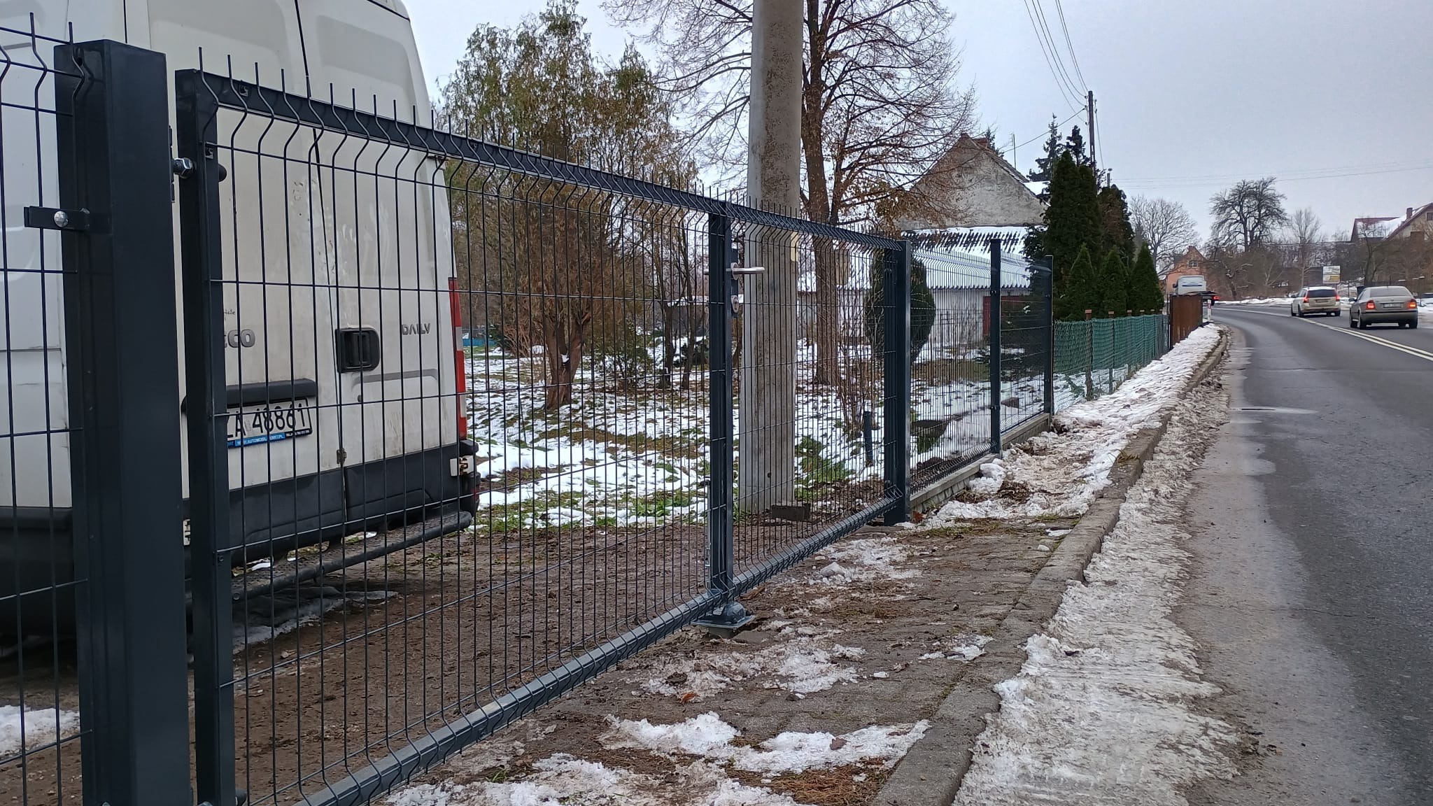 Nowoczesne, grafitowe ogrodzenie z paneli siatkowych, zamontowane wzdłuż chodnika, z częściowo odśnieżonym terenem w tle i zaparkowanym białym samochodem dostawczym po lewej stronie.