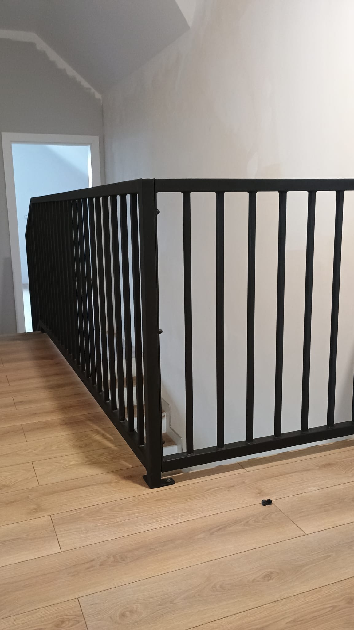 Nowoczesna, czarna balustrada schodowa z pionowych prętów, zamontowana w jasnym wnętrzu z panelami podłogowymi i widocznym fragmentem schodów.