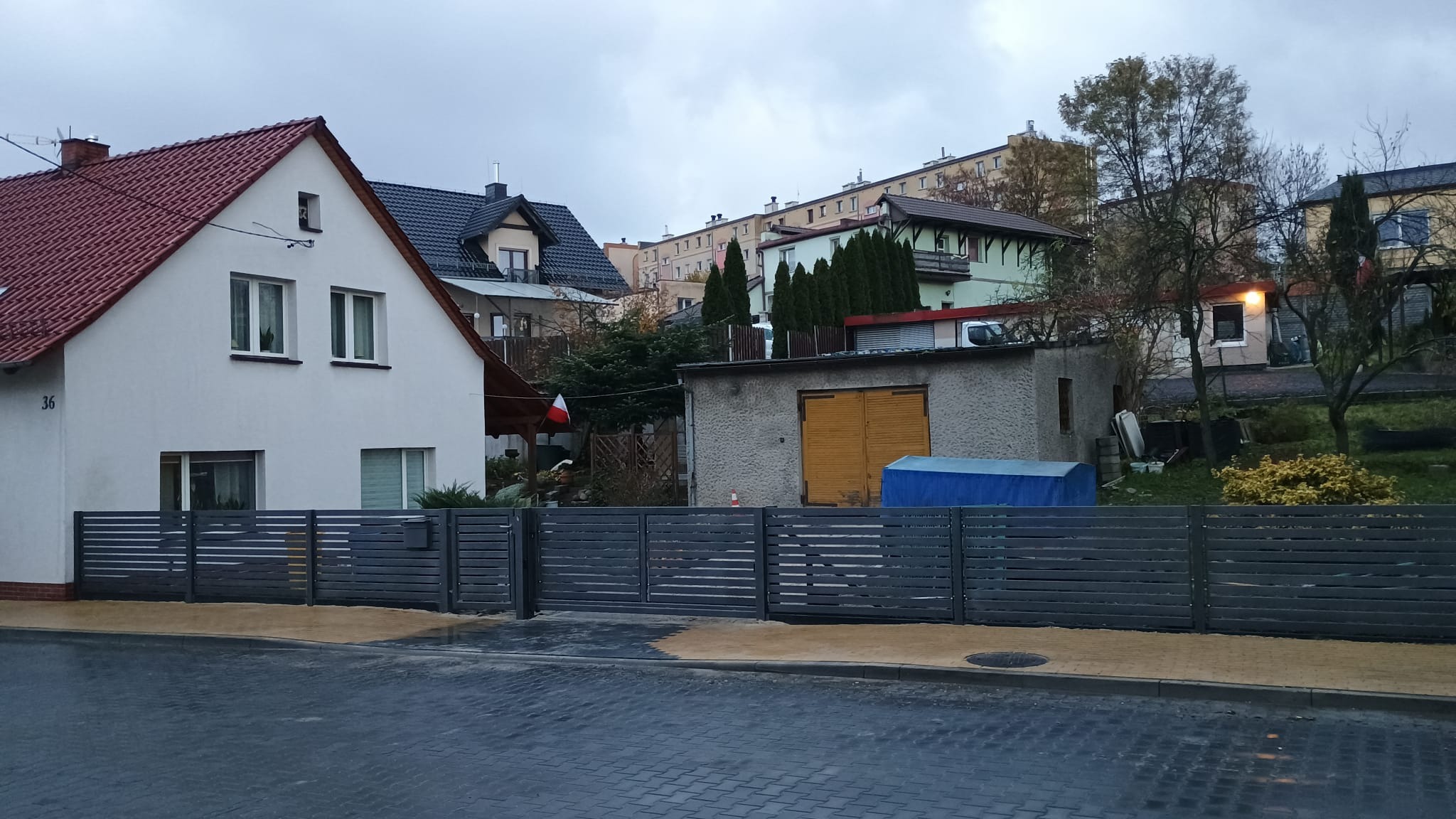 Nowoczesne, poziome przęsła ogrodzeniowe w kolorze grafitowym przed domem z czerwonym dachem, widok na ulicę z kostki brukowej i sąsiednie budynki w tle.