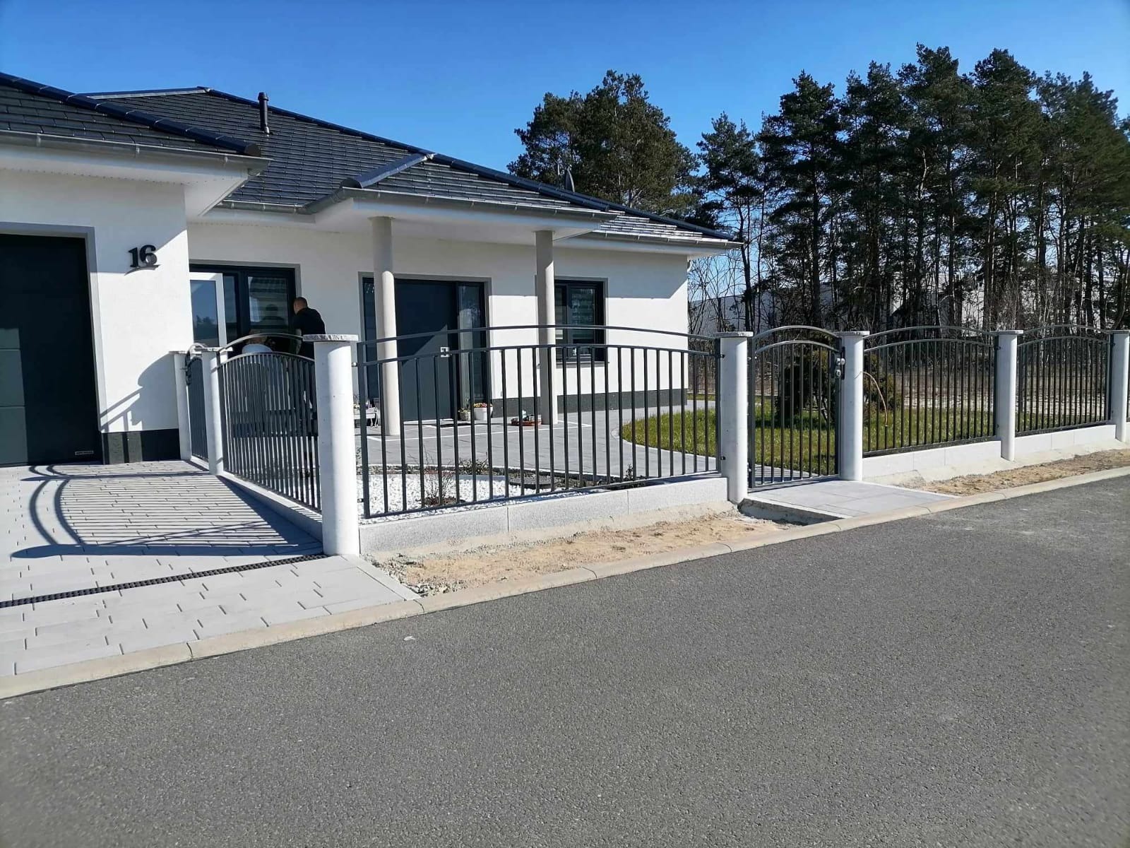 Nowoczesne ogrodzenie z metalowymi prętami i betonowymi słupkami przed białym domem z ciemną dachówką, widok z ulicy w słoneczny dzień.