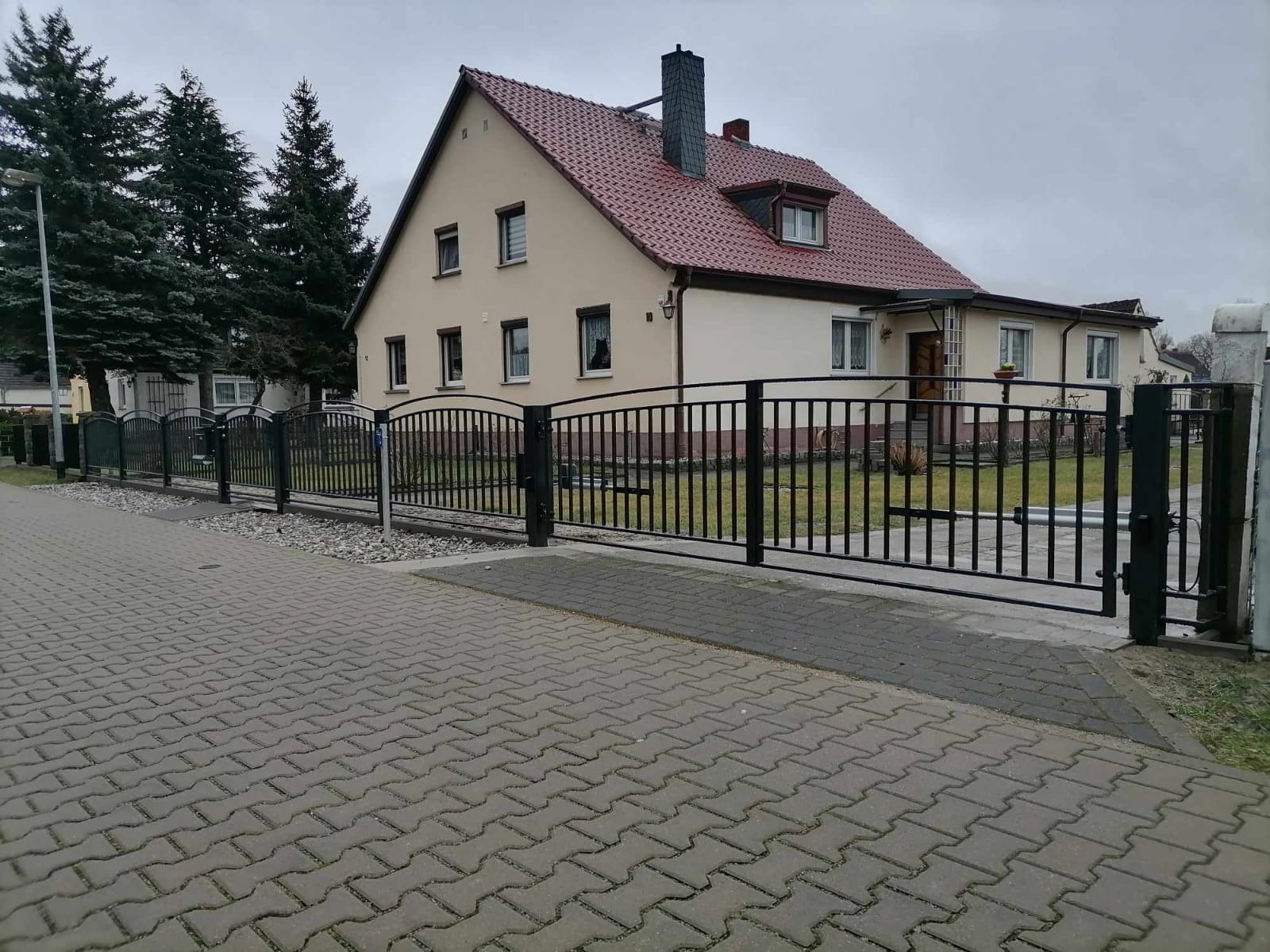 Czarna, metalowa brama skrzydłowa i przęsła ogrodzeniowe przed domem z beżową elewacją i czerwoną dachówką, widok z chodnika wyłożonego kostką.