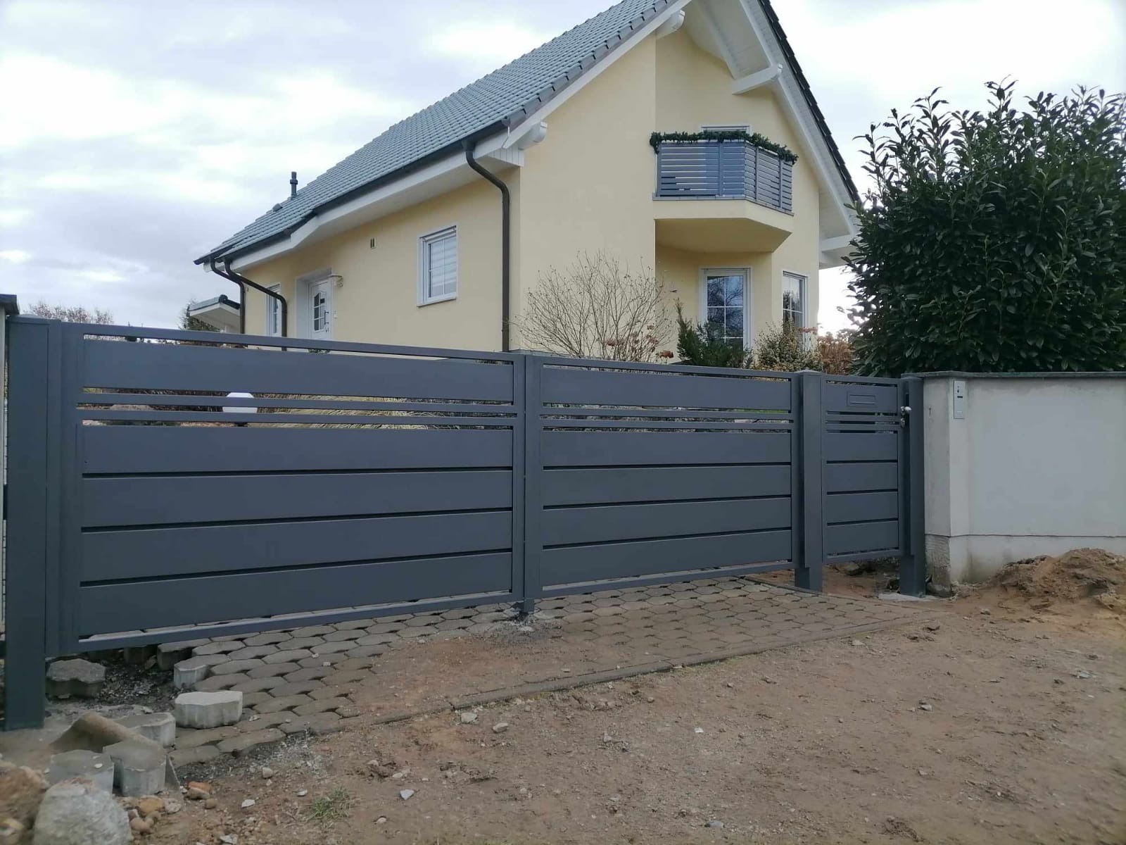 Nowoczesna, szara brama wjazdowa z poziomych paneli, widoczna na tle jasnego domu z balkonem i kostki brukowej.